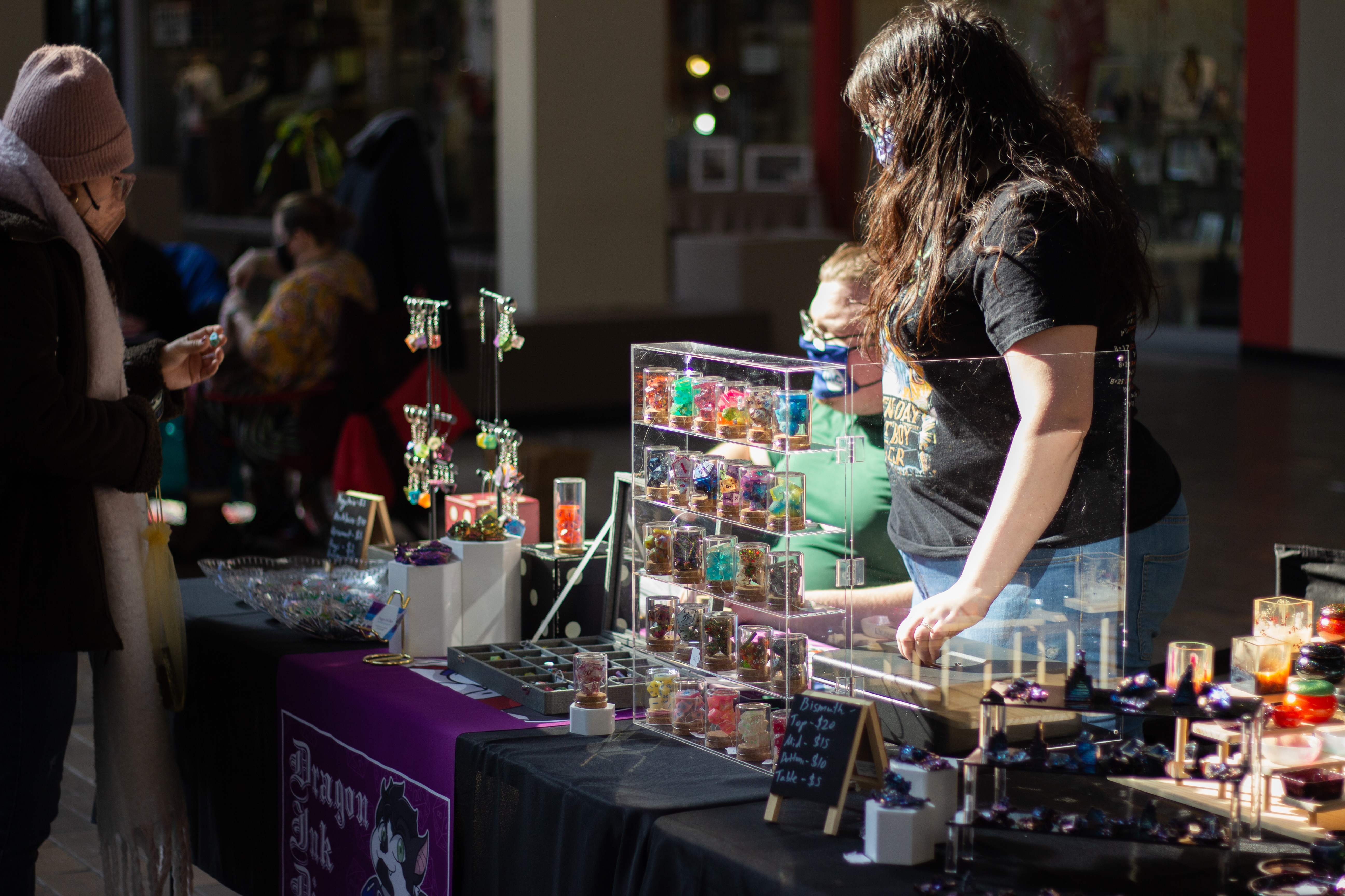 A view of Dragon Ink Diceâ€™s vendor table at the CU Winter Farmers Market located in  Lincoln Square Mall. On the table is an assortment of artisan dice, earrings, dishes and ashtrays of various styles and colors. The table itself is covered in black while the sign for Dragon Ink Dice is purple. The vendor is helping out an interested shopper as they look at one of the dice. Photo by Jorge Murga.