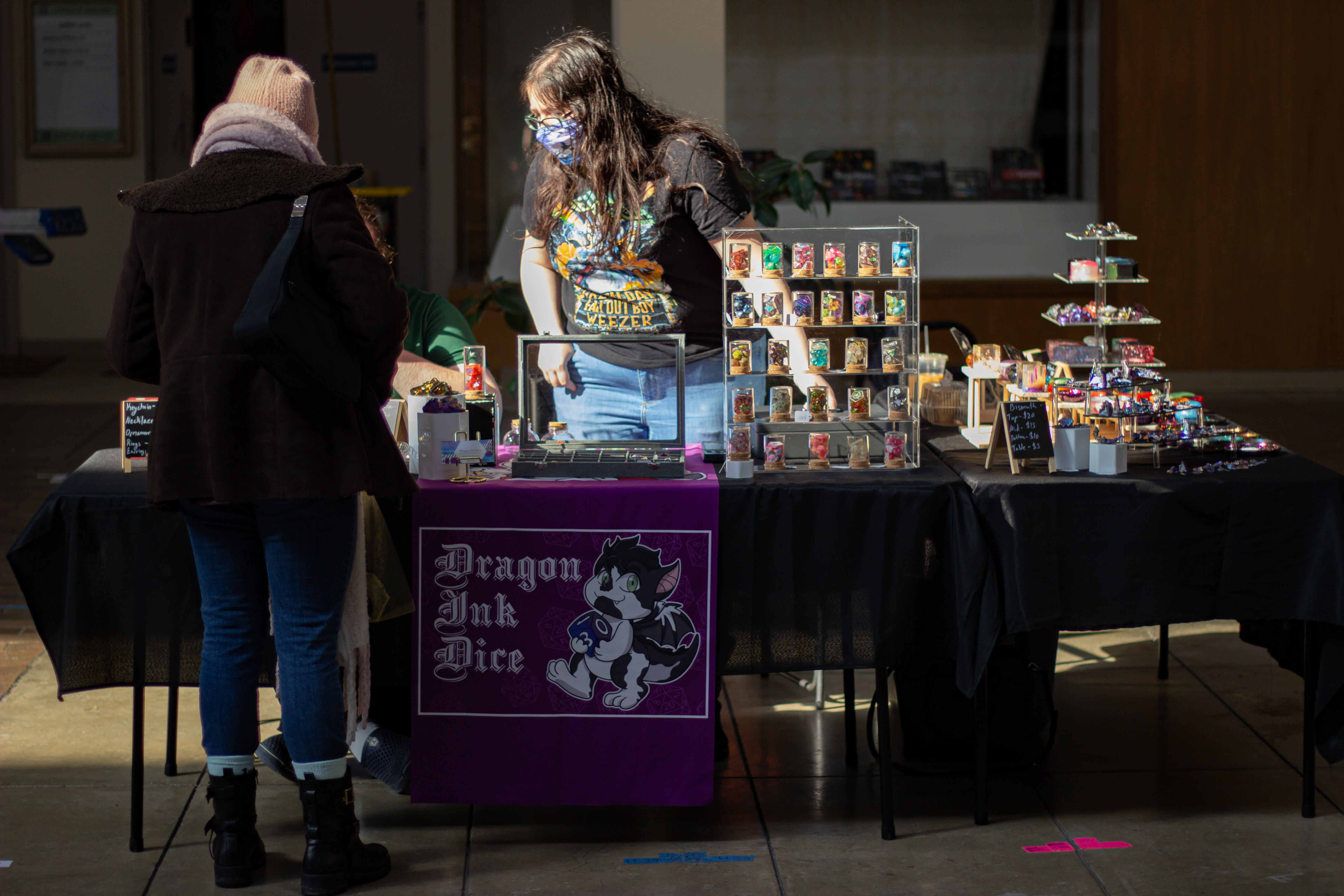 A view directly in front of the Dragon Ink Dice table. The vendor is helping out a shopper. There are an assortment of dice displayed on a shelf. Next to the shelf is a box full of more dice. The right side of the table is where the Bismuth, ashtrays, jewelry boxes, and other accessories are displayed. Photo by Jorge Murga.