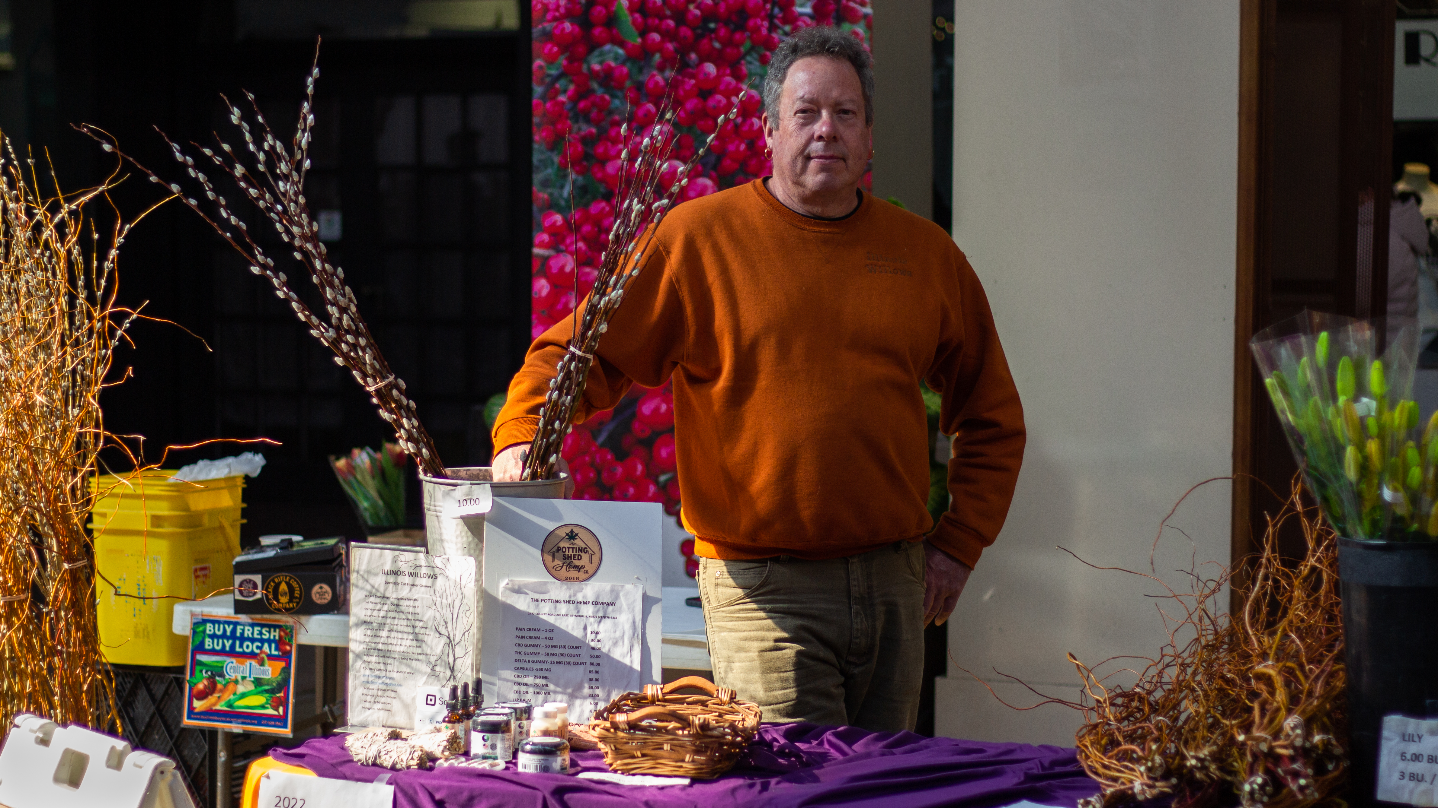 In view of the stand for Illinois Willows at the CU Winter Farmers Market located in Lincoln Square Mall. The table has a purple cloth over it and has an array of flowers, sage, and an assortment of CBD products. The representative of Illinois Willows, Kent Miles, stands behind the products. There is also a sign that reads â€œThe Potting Shed Hemp Companyâ€ and it lists the   prices for the different CBD products. Photo by Jorge Murga.
