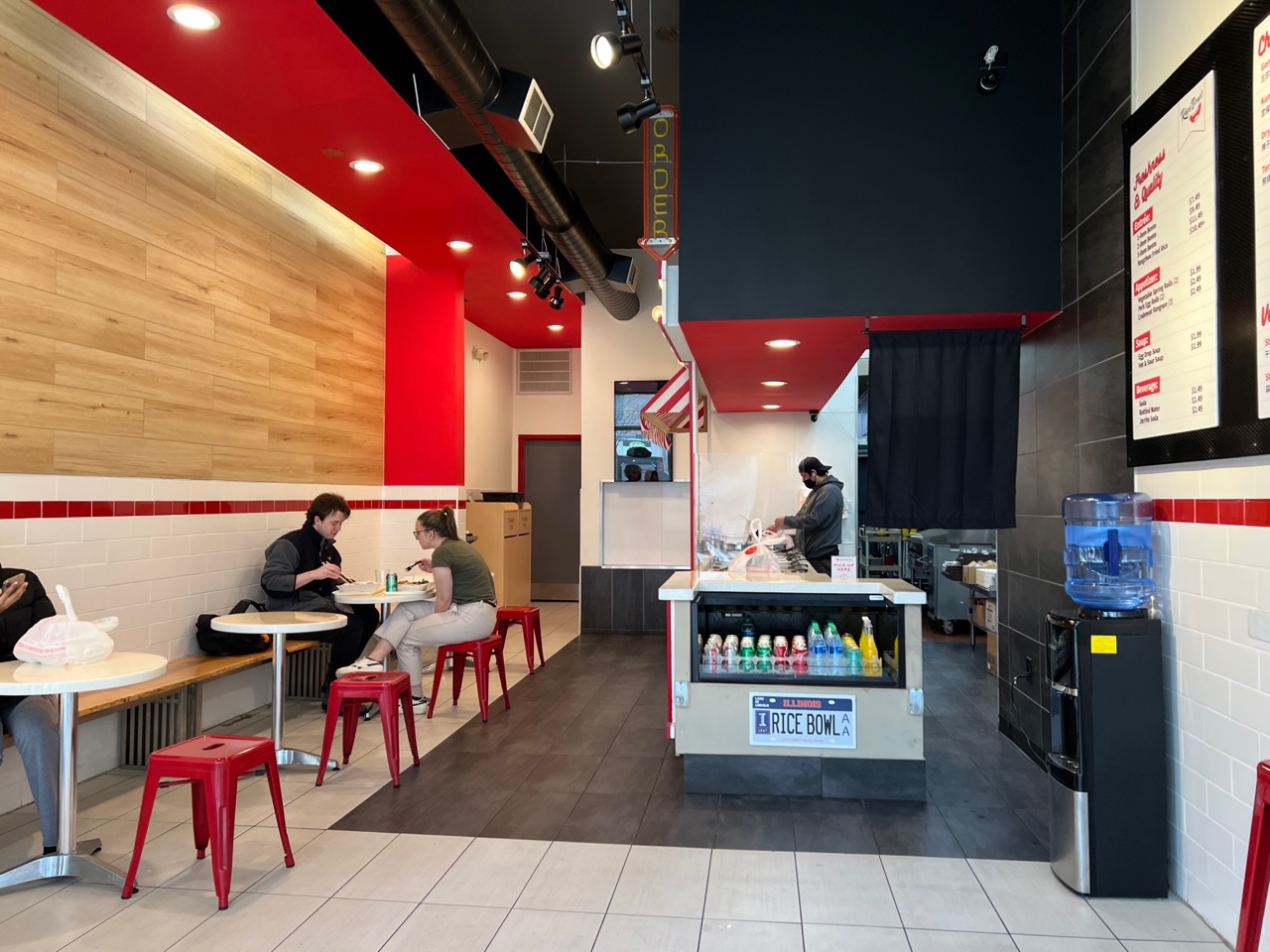The interior of Rice Bowl has a red ceiling and several circle tables along the left wall. There is a counter in the middle that has a single employee in a black shirt preparing food for the line. Photo by Alyssa Buckley.