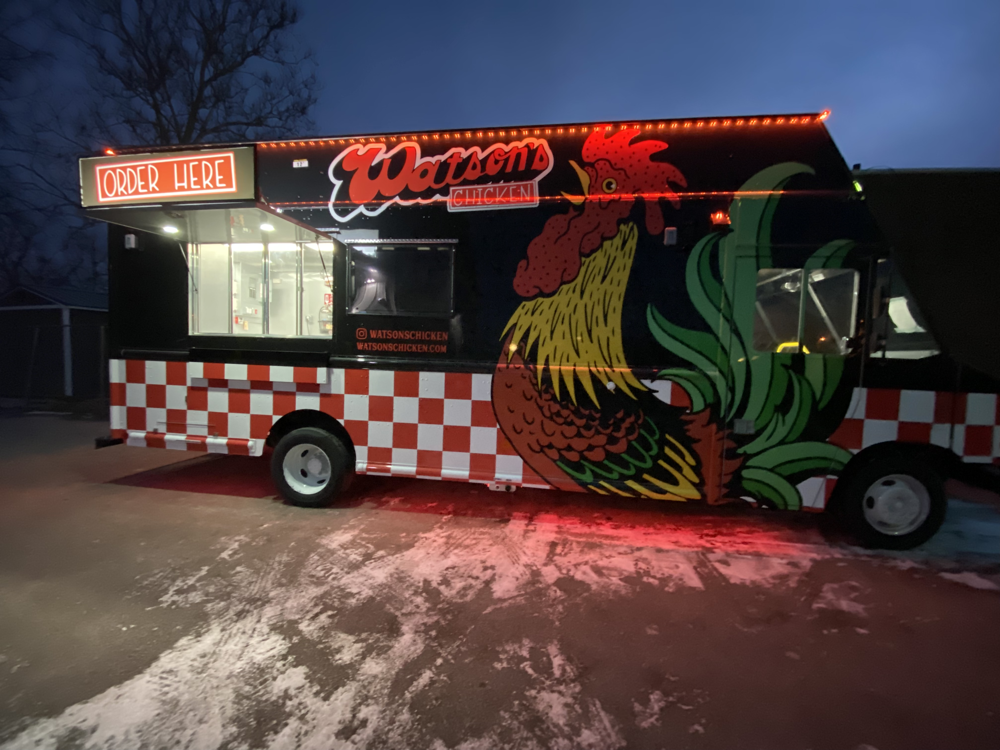 A Watson's food truck painted black with a white and red checked bottom, with a large illustration of a chicken on the side, featuring a walk up window.