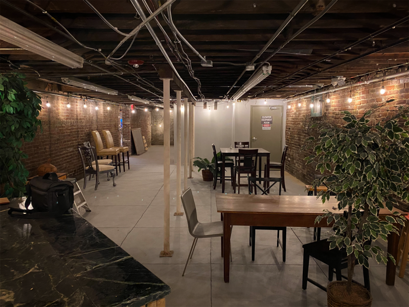 A large basement room with exposed brick walls. String lights run along the ceiling, and there are a few sets of tables and chairs scattered throughout. Photo by Andy Long. 