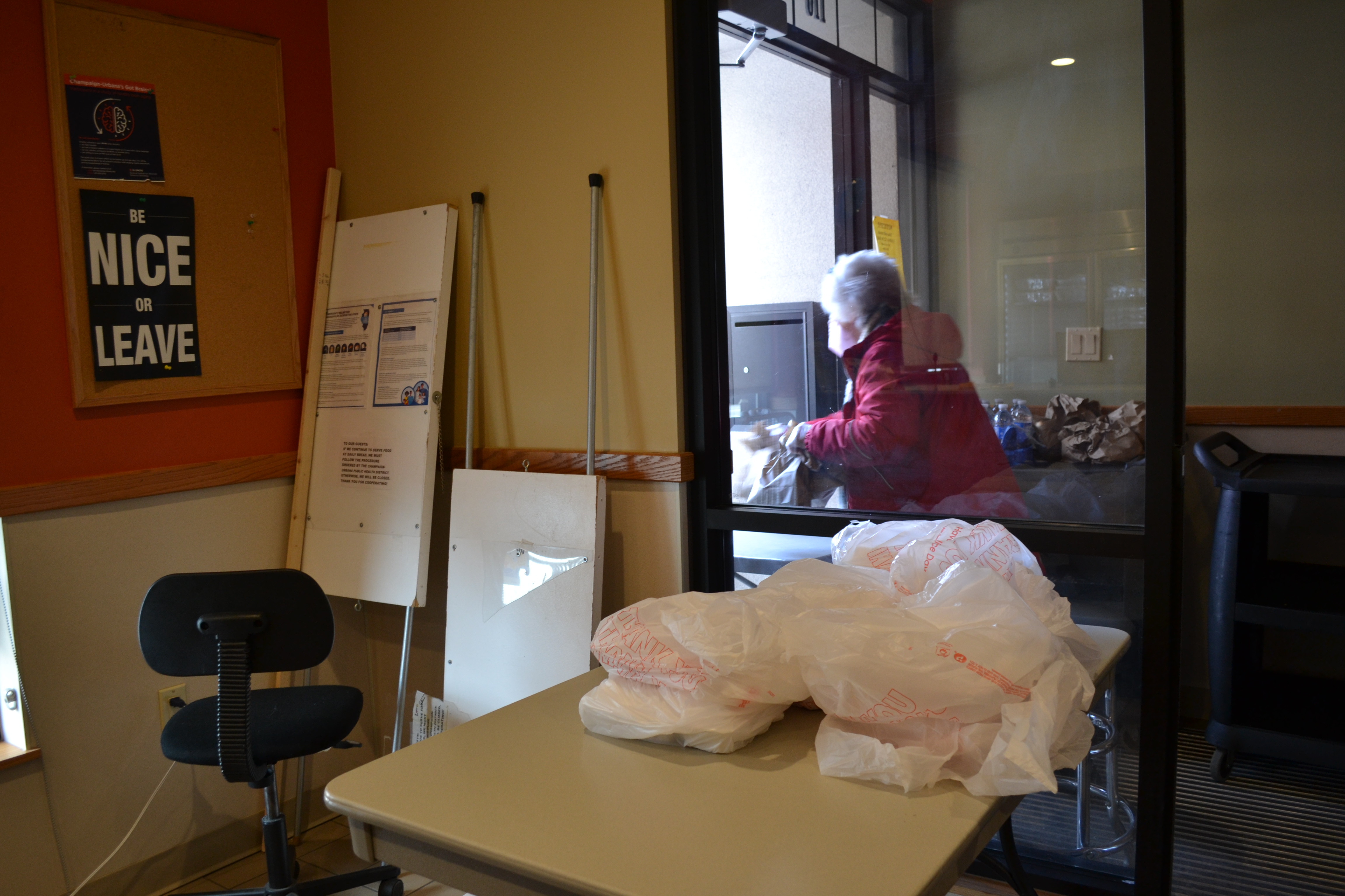 At the Daily Bread Soup Kitchen, there is a folding table with lunches ready to be distributed. In a small glass room, the door is open to First Street. Ruth Ann and Charlie Evans are masked and giving out meals. Photo by Alyssa Buckley.
