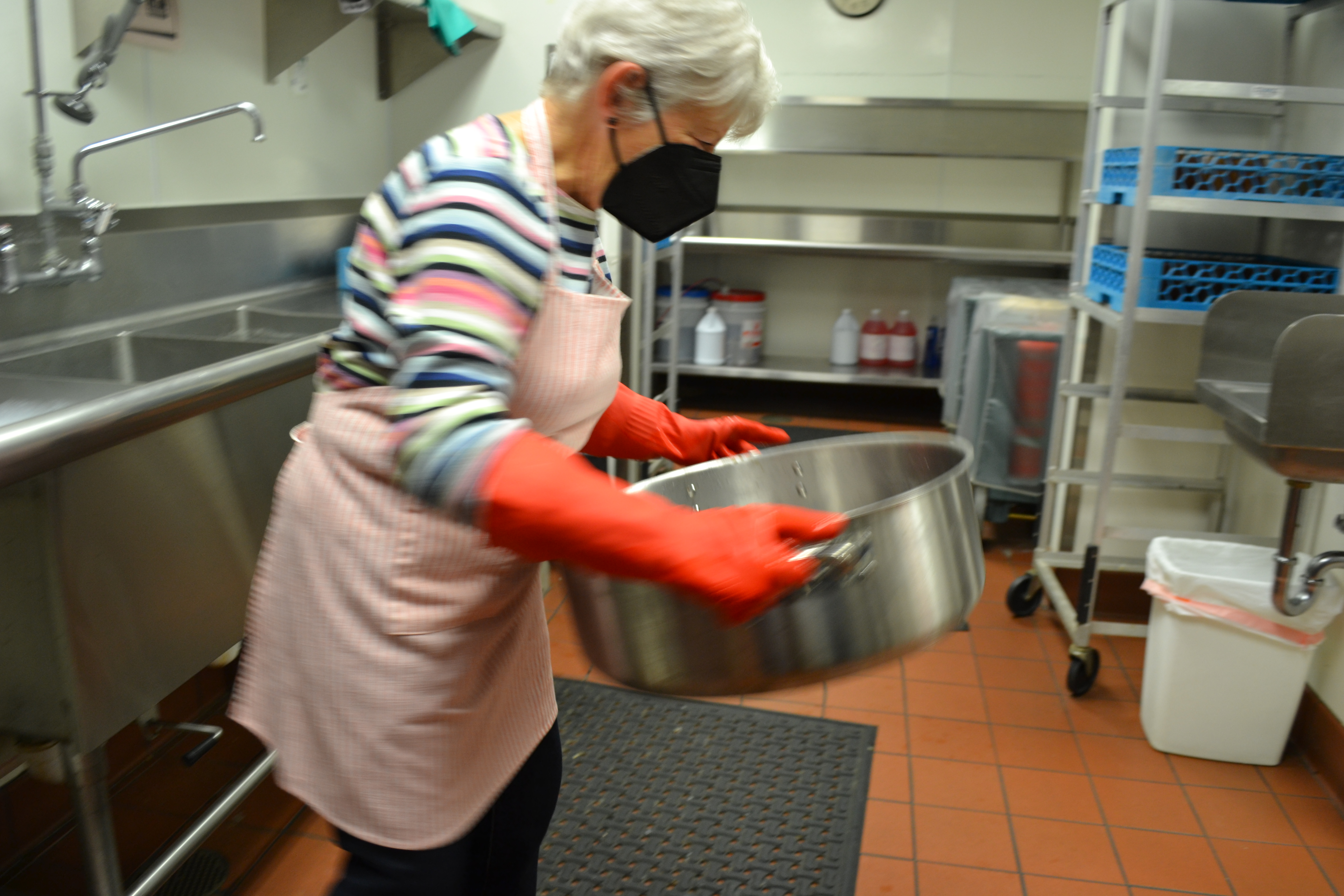 A gray-haired, masked volunteer carries a heavy pot in the dishwashing room. Photo by Alyssa Buckley.