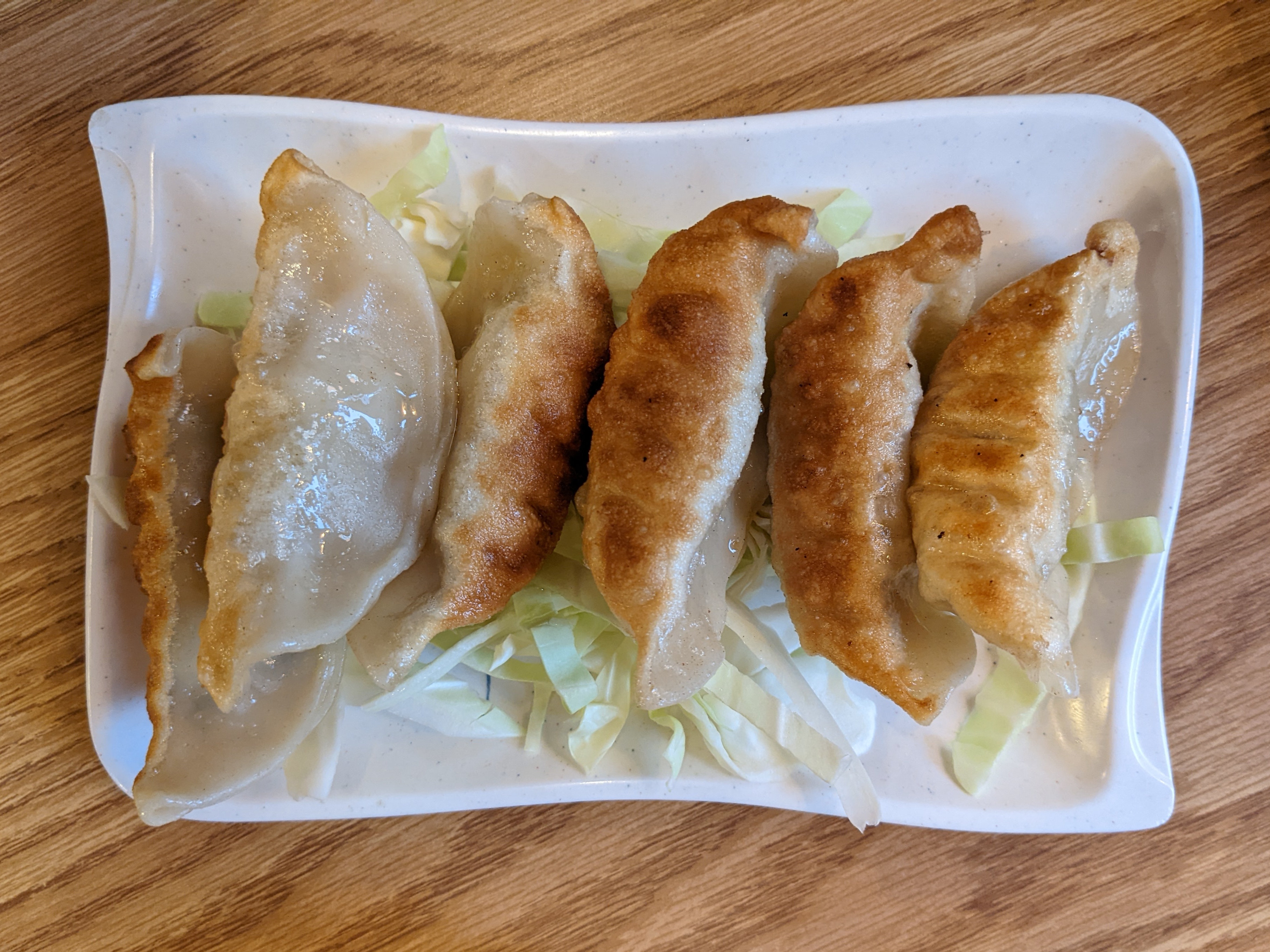 On a wooden table, there is a white plate with six fried dumplings on a bed of cabbage. Photo by Tayler Neumann.