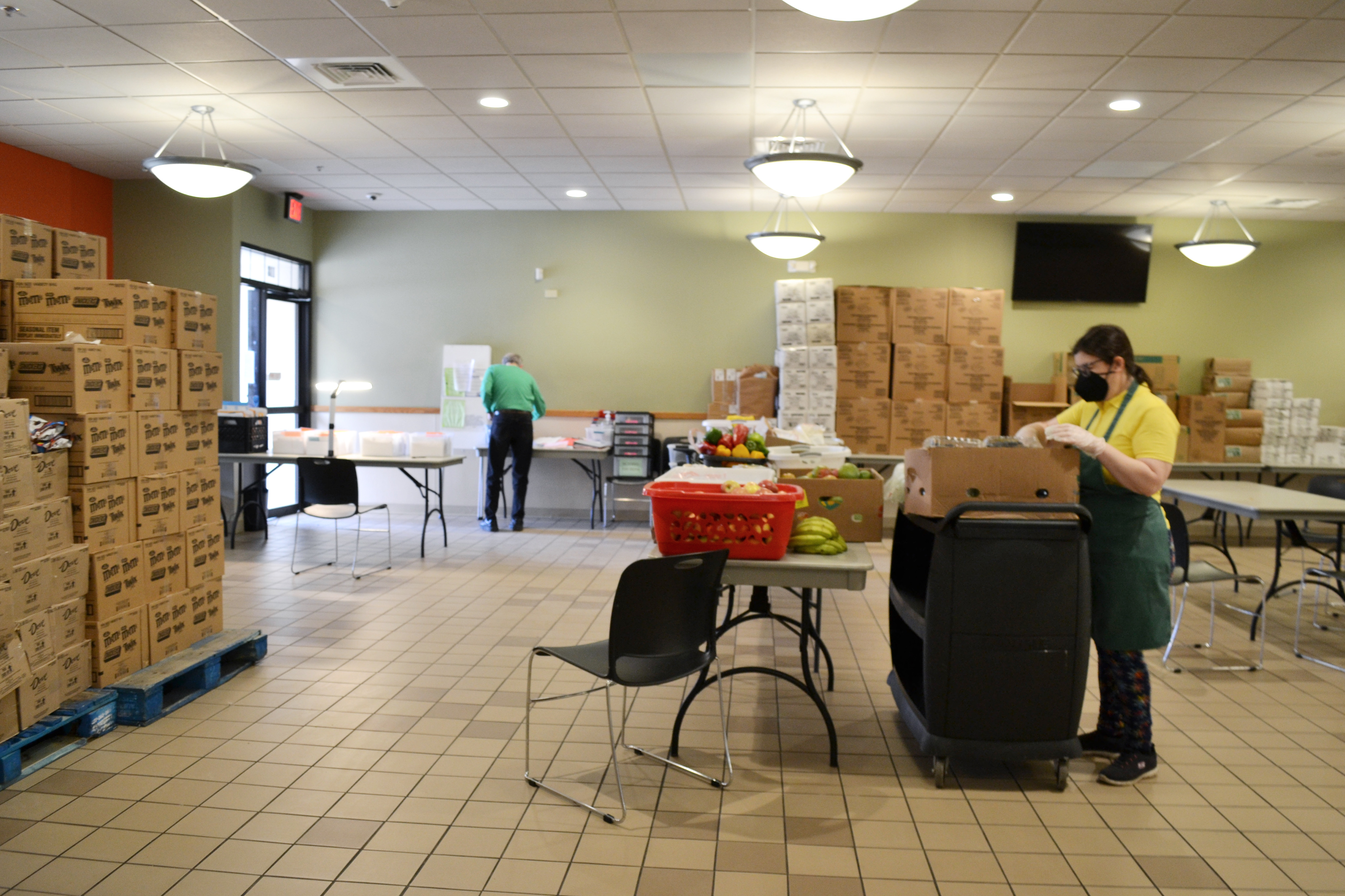 Inside the Daily Bread Soup Kitchen, there are volunteers preparing for lunch. On the left there are brown boxes of donated food stacked, and folding tables with produce and more boxes. Photo by Alyssa Buckley.