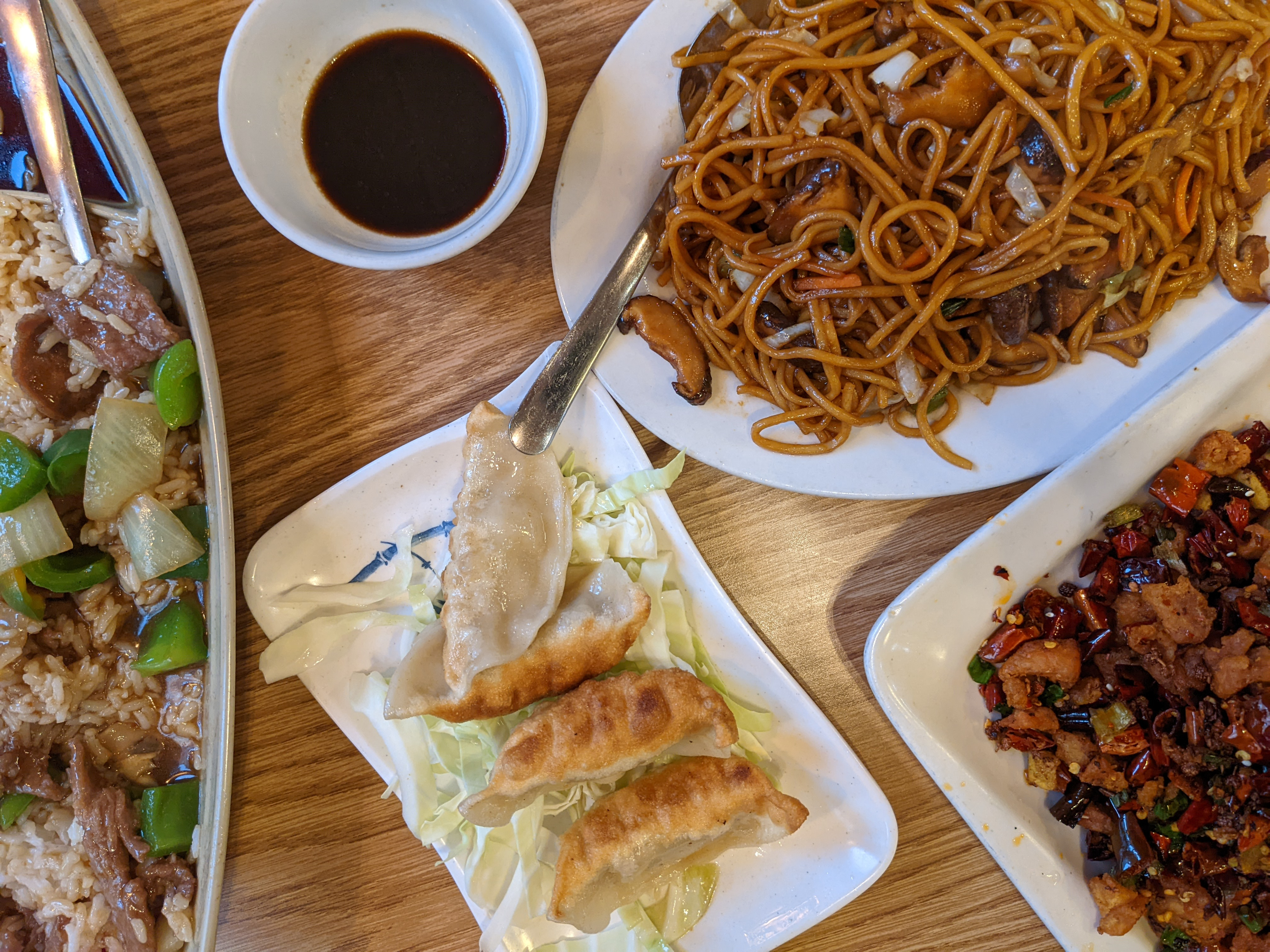 On a wooden table, there are four dishes and one cup of dark sauce. There are beef and veggies in a long dish on the left, dumplings on a plate in the center bottom, and the top right has noodles with the bottom right dry chili chicken. Photo by Tayler Neumann.