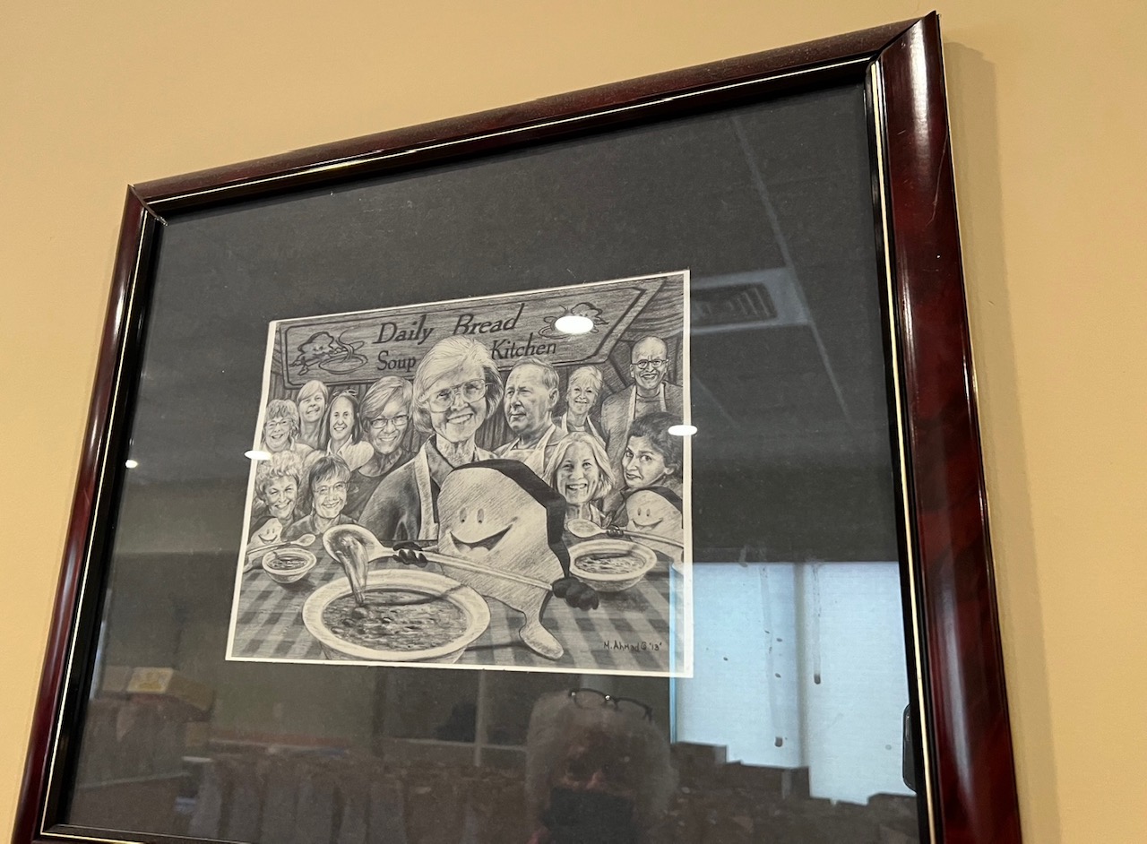 On a yellow wall, a framed drawing of Ellen McDowell with other Daily Bread Soup Kitchen people. In the reflection, you can see Ruth Ann Evans, the interviewee. Photo by Alyssa Buckley.