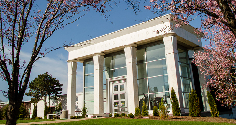 The fifth street entrance of Krannert Art Museum in spring. The front of the building has a white roof and four white columns that stand in front of an all glass facade. There are flowering trees on either side. Photo from Krannert Art Museum Facebook page.