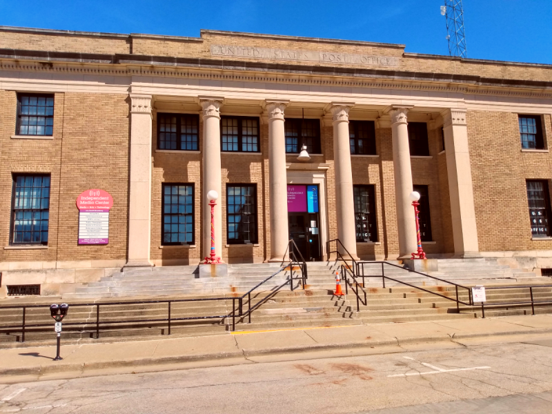 An external shot of the Independent Media Center in the old post office building in downtown Urbana. It is a brick facade with columns in the neoclassical style. The entrance is above street level and is accessed by a steps guarded by railings. A multicolored sign on the left indicates the current tenants, including the IMC. Photo by Michael O'Boyle.