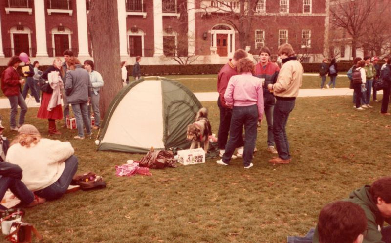 A photo from 1984, groups of students are gathered on the Quad. In the center is a small white and green tent with a dog and a case of beer next to it. Photo from Champaign Urbana History Facebook group.