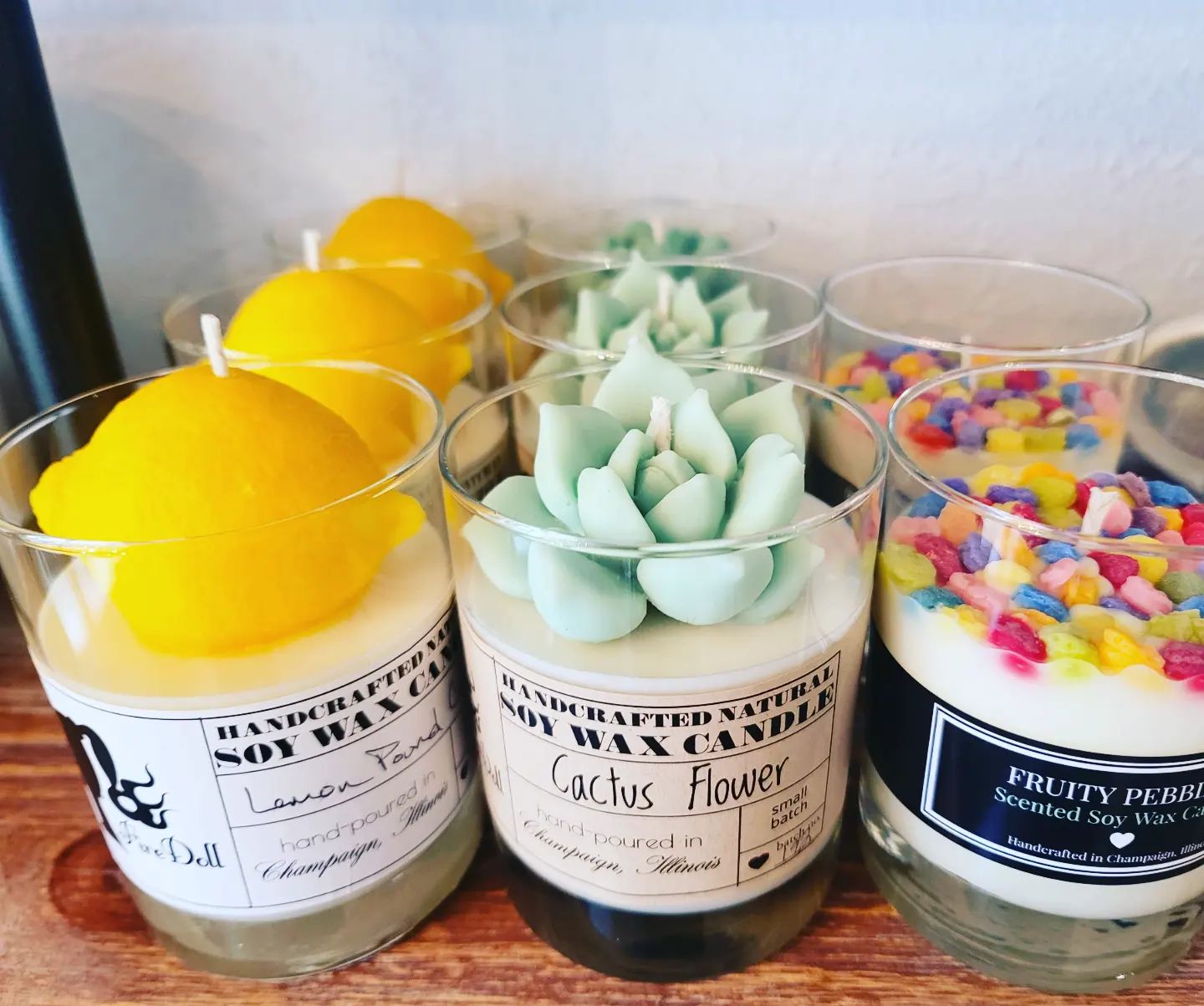 Three candles in glass jars on a shelf. On the left, a candle with a big yellow lemon on top; in the middle a candle with light green cactus flowers on top; on the right, a candle with fruity pebbles cereal on top. Photo from the Firedoll Studio Facebook page.