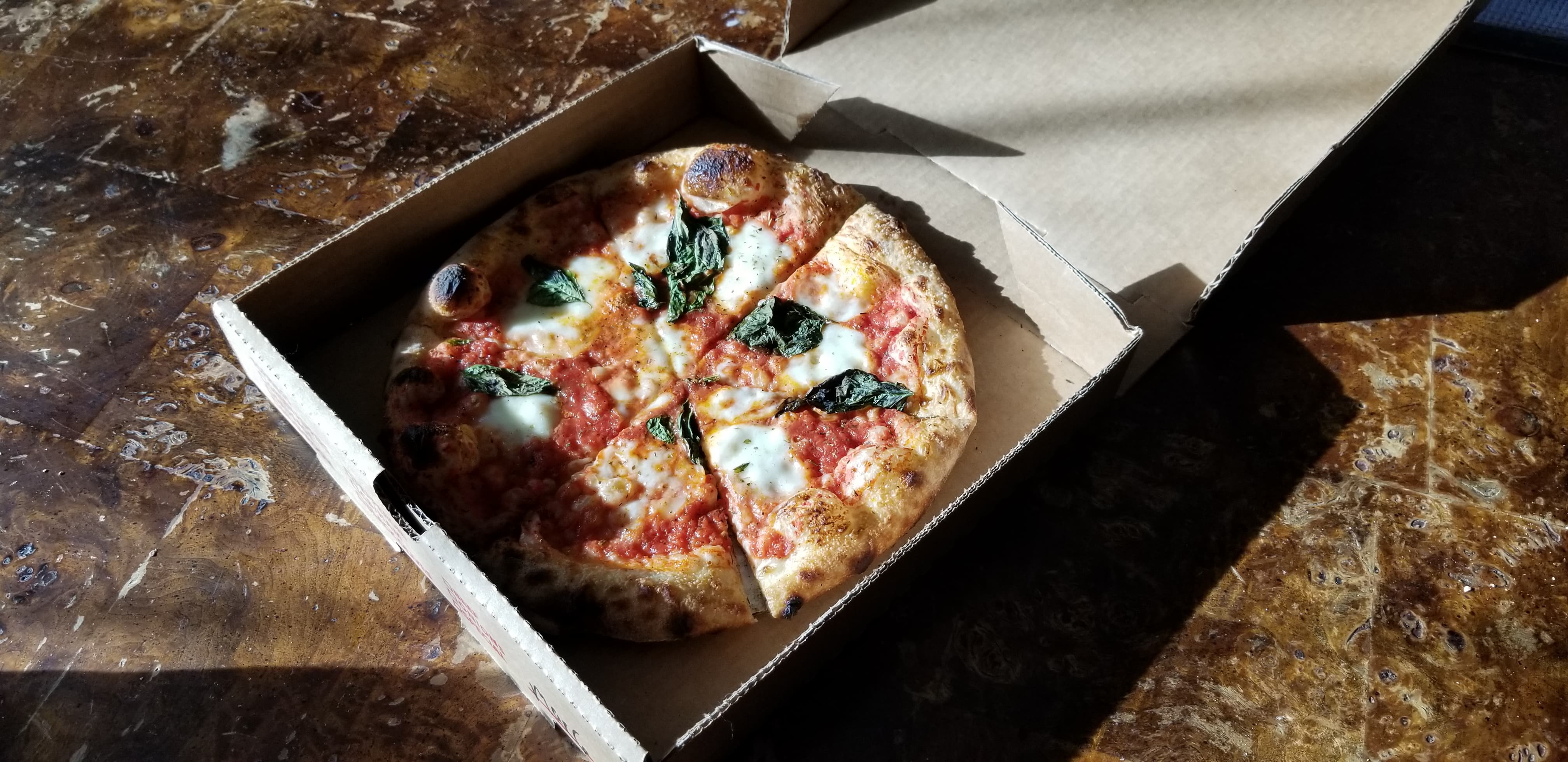 On a sunny brown table, there is a pizza in a paper box. Photo by Nina Hopkins.
