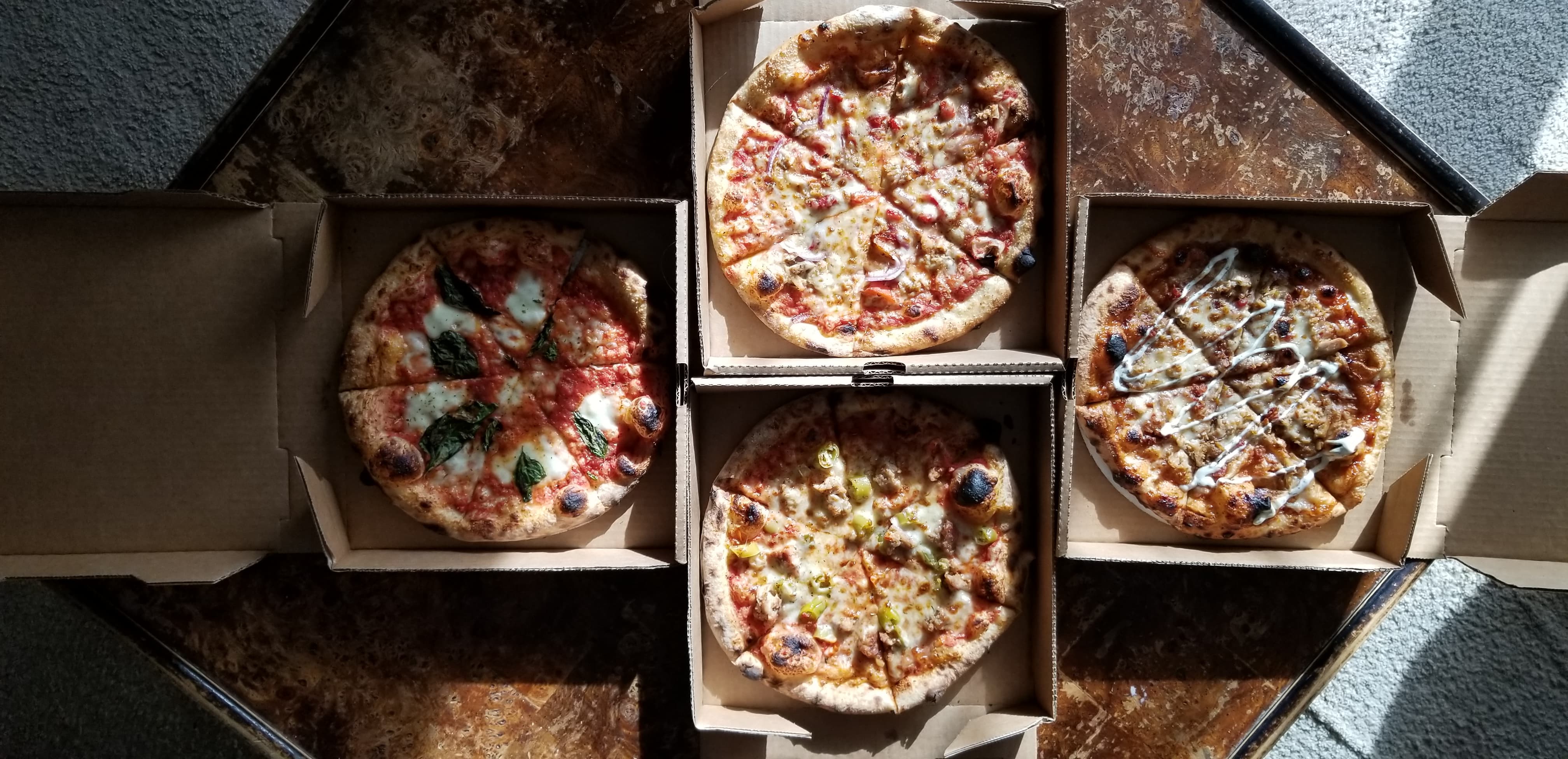 On a brown table, there are four pizzas in cardboard pizza boxes. The boxes are all open, and the sliced full pizza pies are circles inside the brown square boxes. Photo by Nina Hopkins.