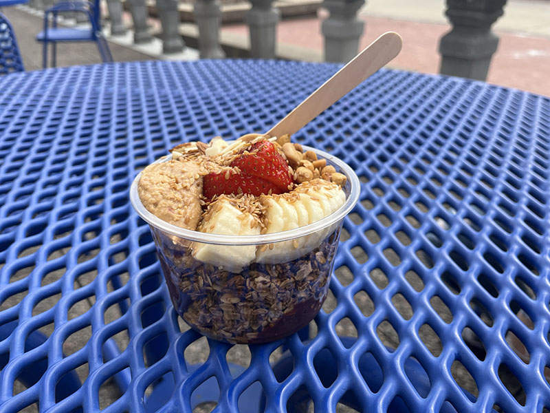 A clear plastic bowl sits on a blue mesh picnic table. Layers of acai and overnight oats fill the bottom of the bowl. Banana slices, peanut butter, peanuts, and strawberries are visible on top. A wooden spoon sticks out to the side. Photo by Julia Freeman.