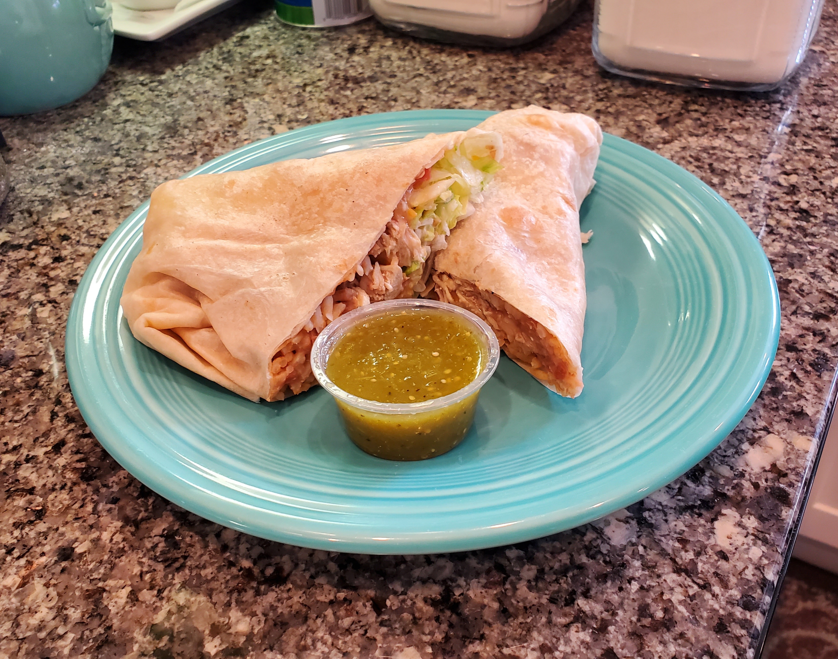 On a light blue plate, there is a chicken burrito cut in half with a small cup of verde salsa in front. Photo by Carl Busch.