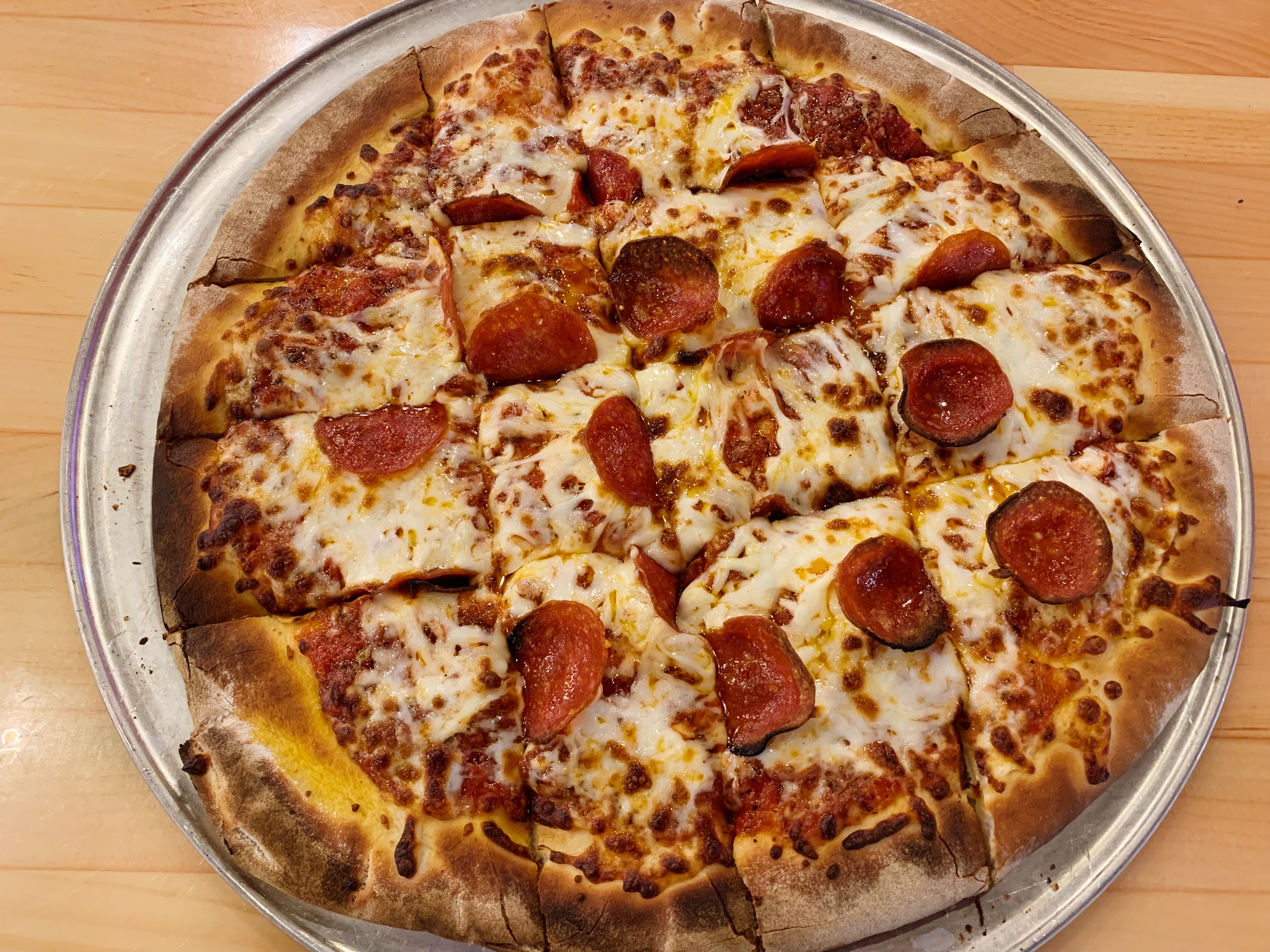 On a wooden table, there is a silver metal circular tray holding a cut but untouched pan of pepperoni pizza. Photo by Anya Uppal.