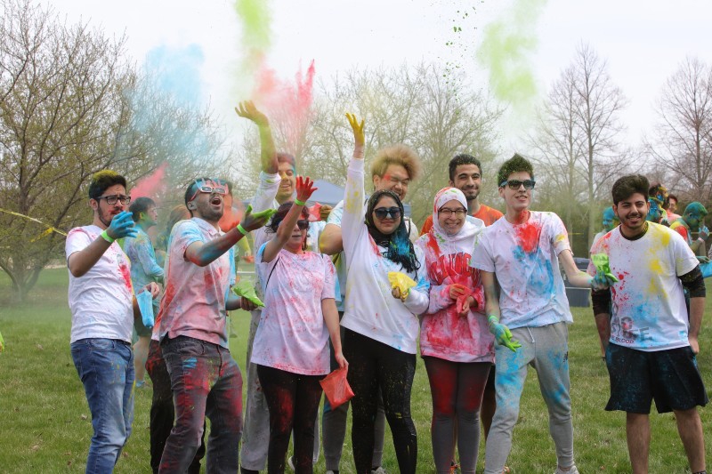 A group of college students are gathered in a group. They are wearing white shirts that are covered in different colors of powder. They are throwing colored powder into the air. Photo from Facebook event page.