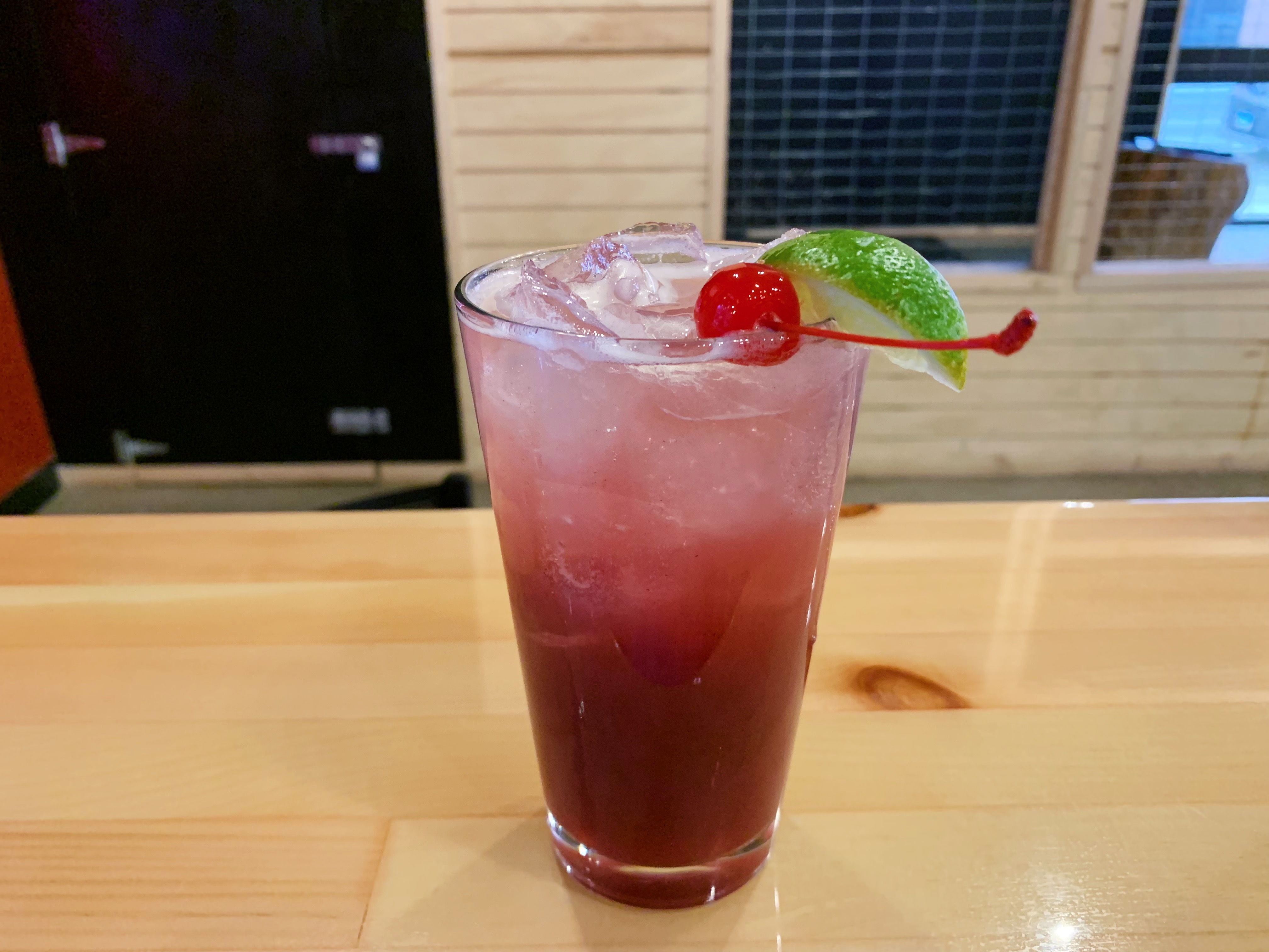 On a wooden table, there is a red drink in a pint glass with a cherry on top of the ice. Photo by Anya Uppal.