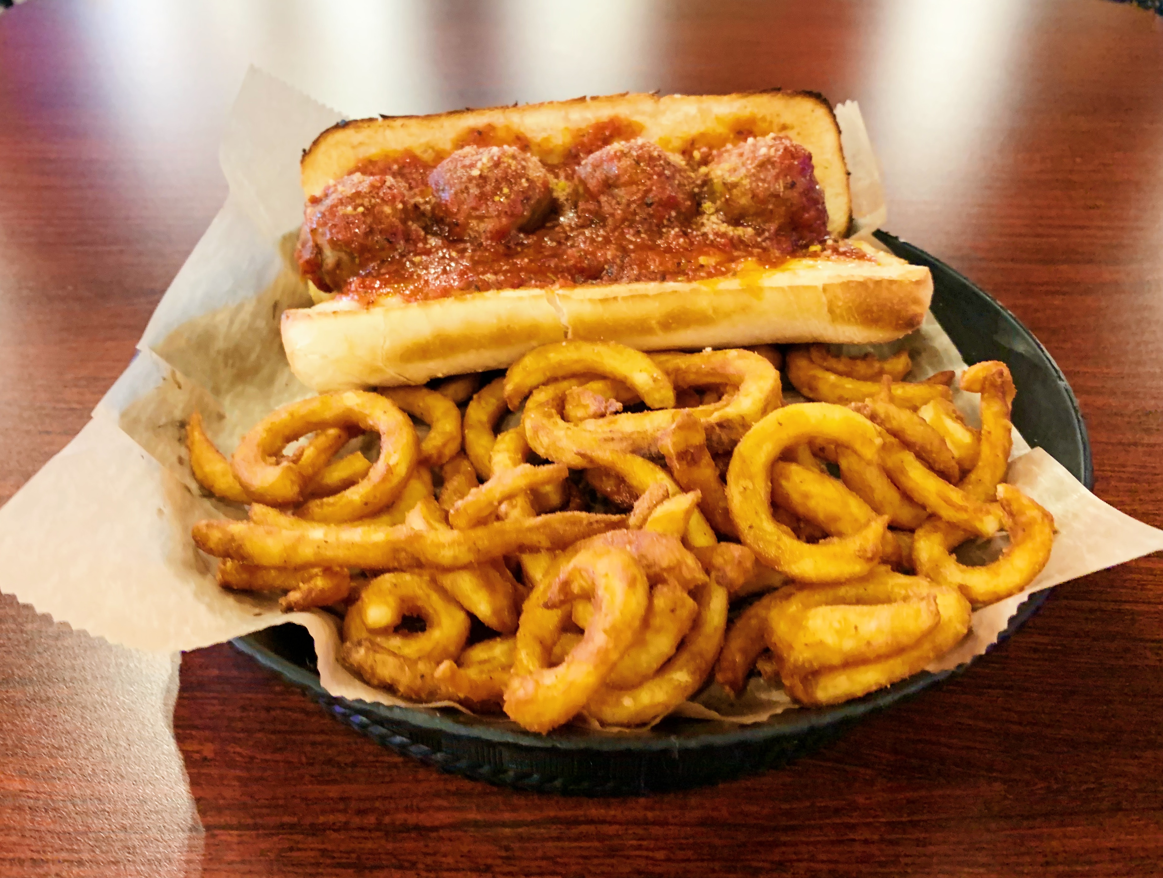 On a wooden table, there is a black basket lined with parchment paper and full of curly fries and a meatball sub. Photo by Anya Uppal.