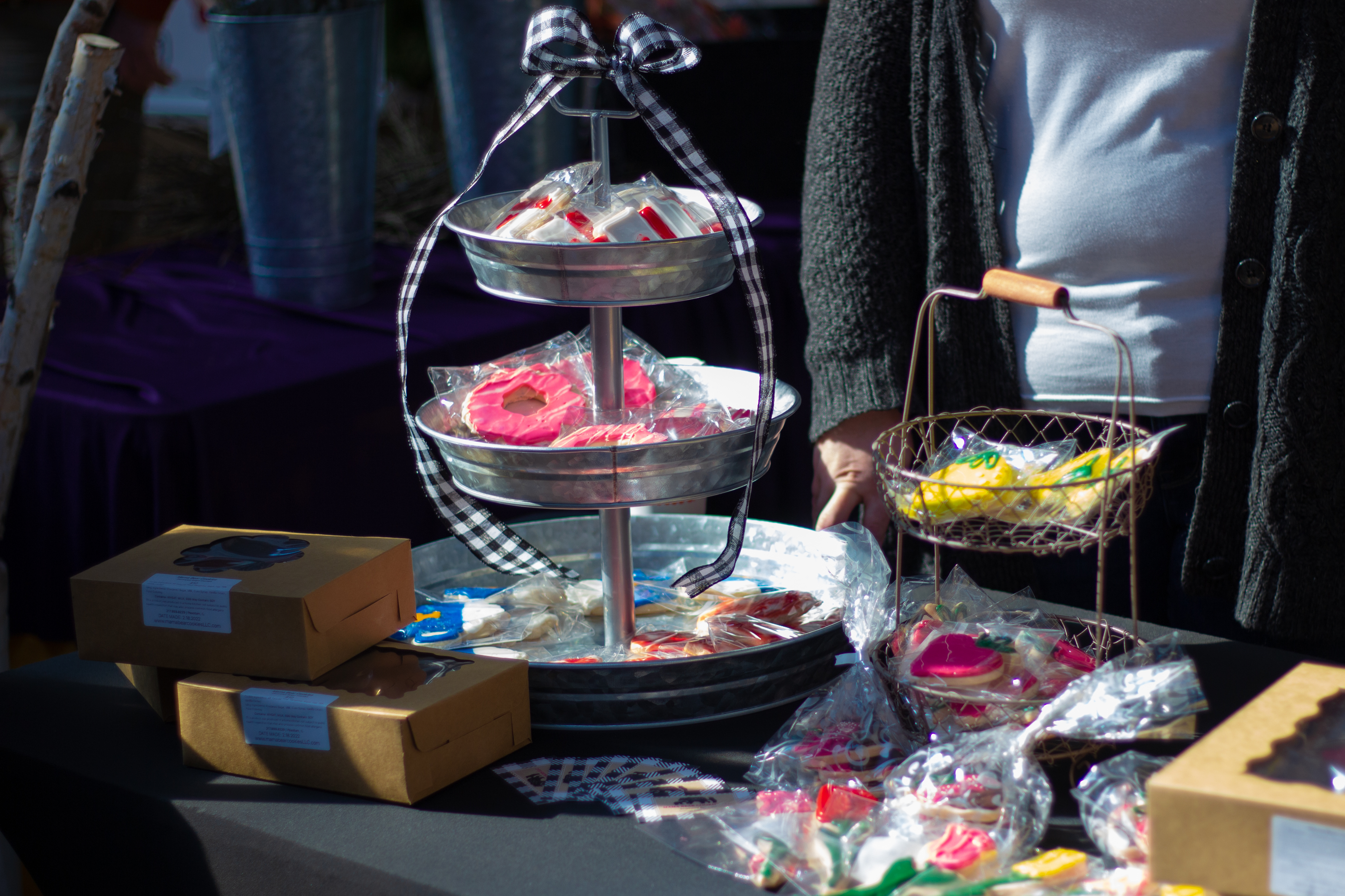 At Urbana's indoor Winter Farmers' Market, a vendor stands behind her desserts for sale. There is a metal tiered display of cookies on a black tableclothed table. Photo by Jorge Murga.