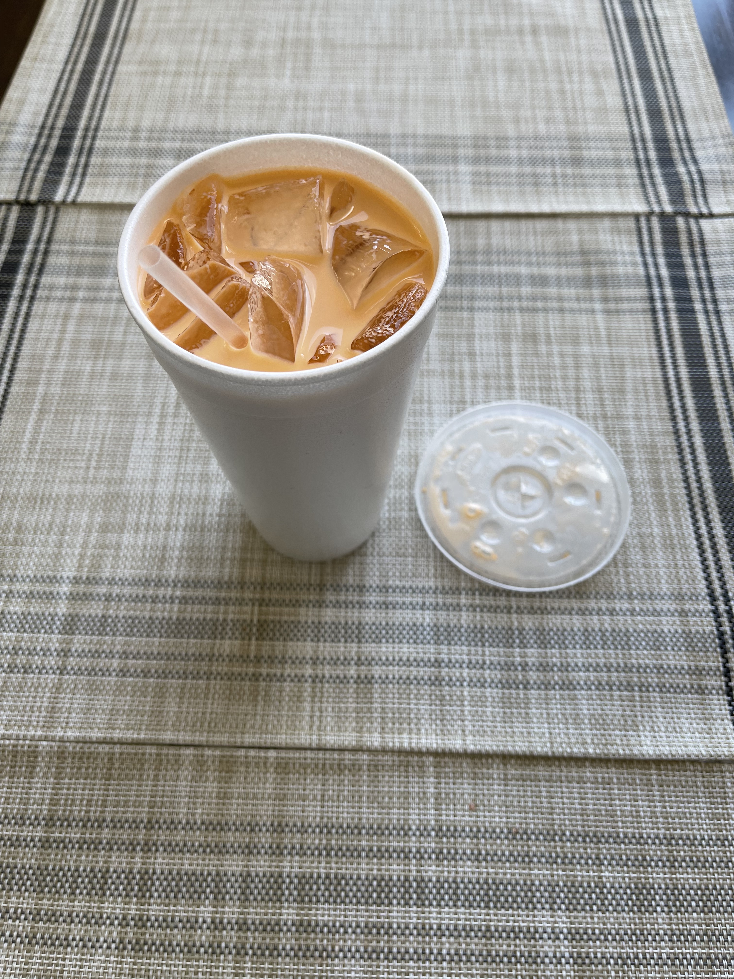 On a brown tablecloth, there is a to go cup with the cover removed. Inside, there is a light brown Thai iced tea with ice. Photo by Shrivatsa Ravikumar.