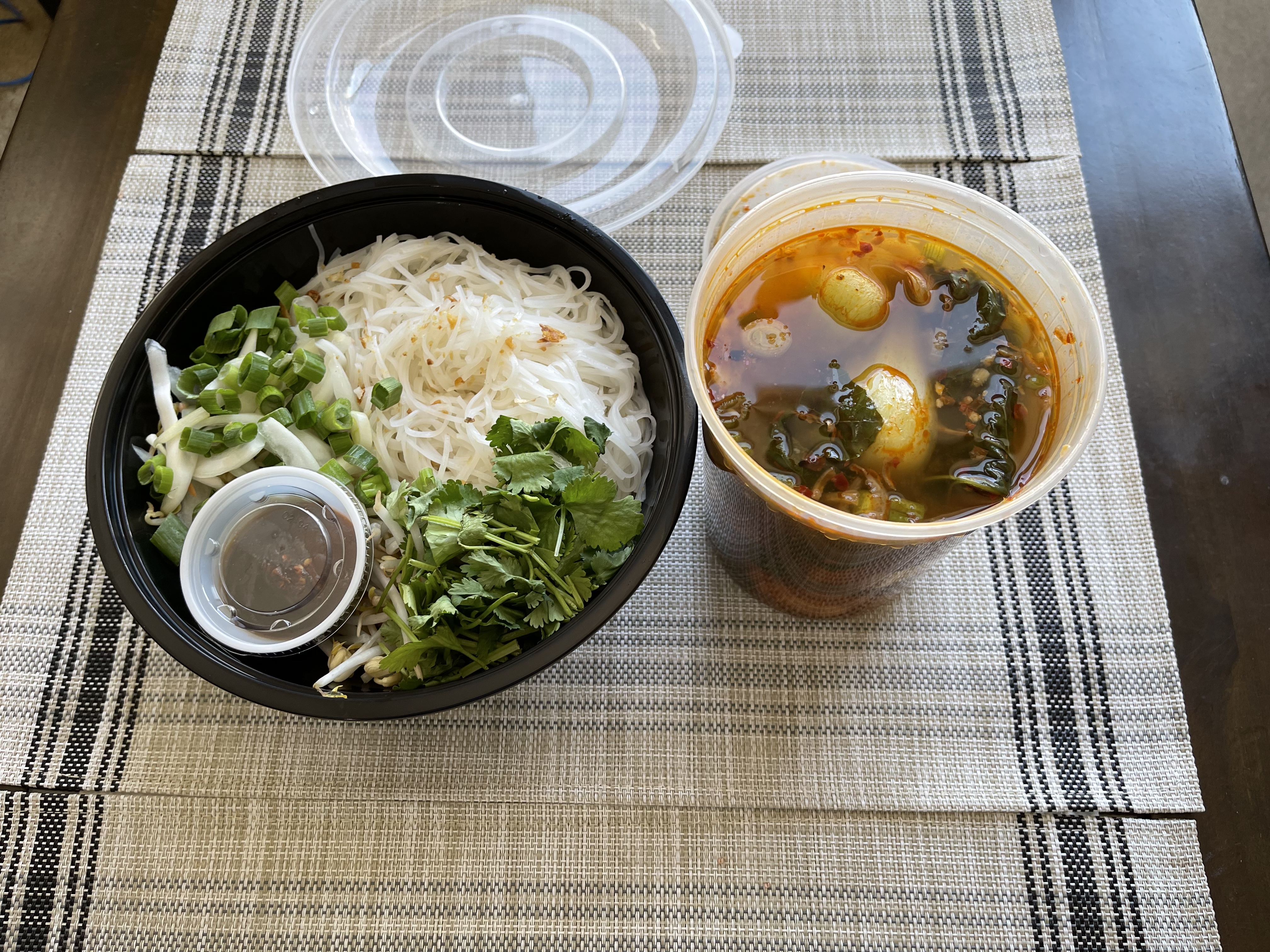 On a brown table with a light brown tablecloth, there is a black bowl of pho broth with scallions, sauce, and noodles. Beside it, there is another takeout container. Photo by Shrivatsa Ravikumar.