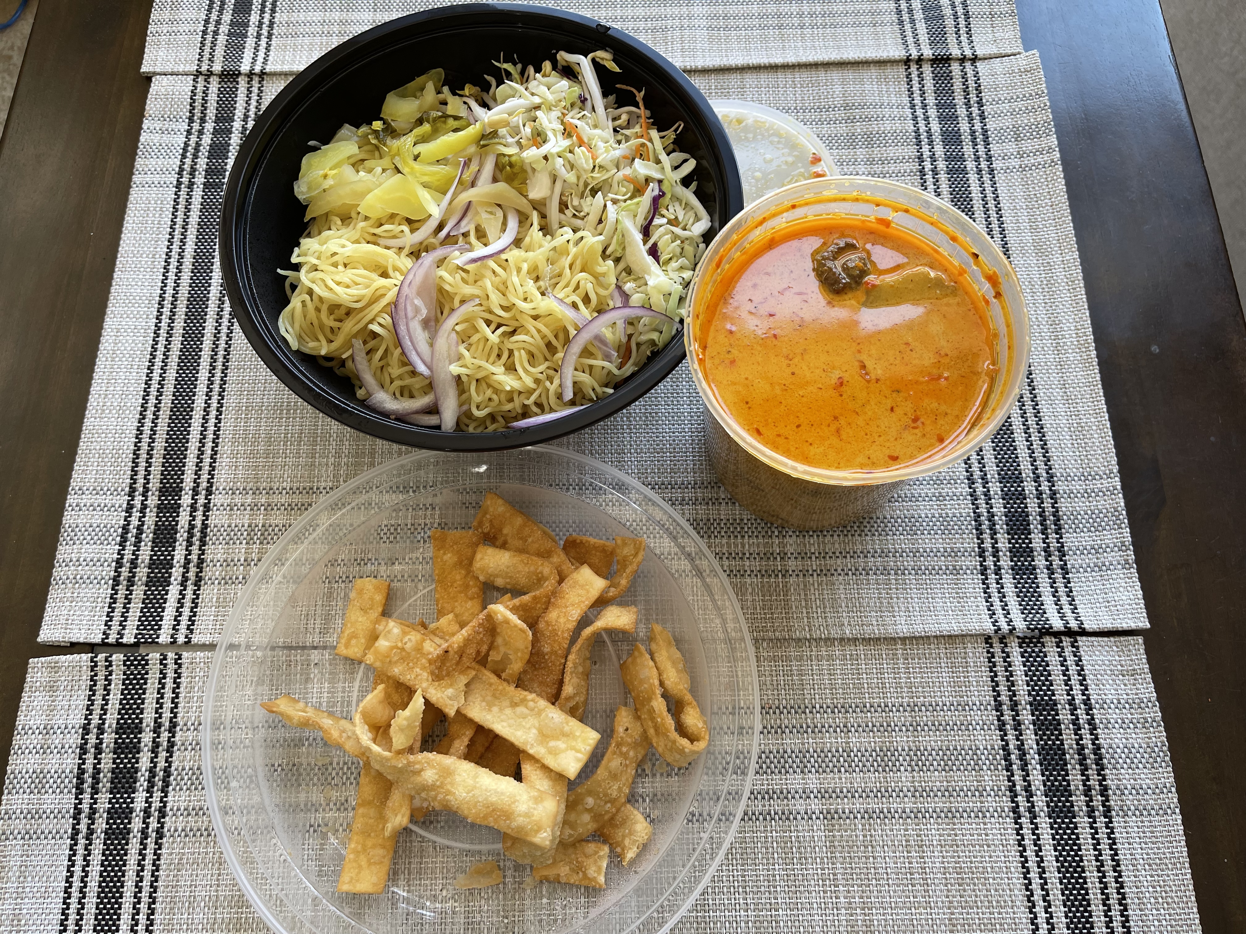 On a table with a brown tablecloth, there are three containers. One is a yellow curry, one is crunchy sticks, and another has noodles and toppings. Photo by Shrivatsa Ravikumar.