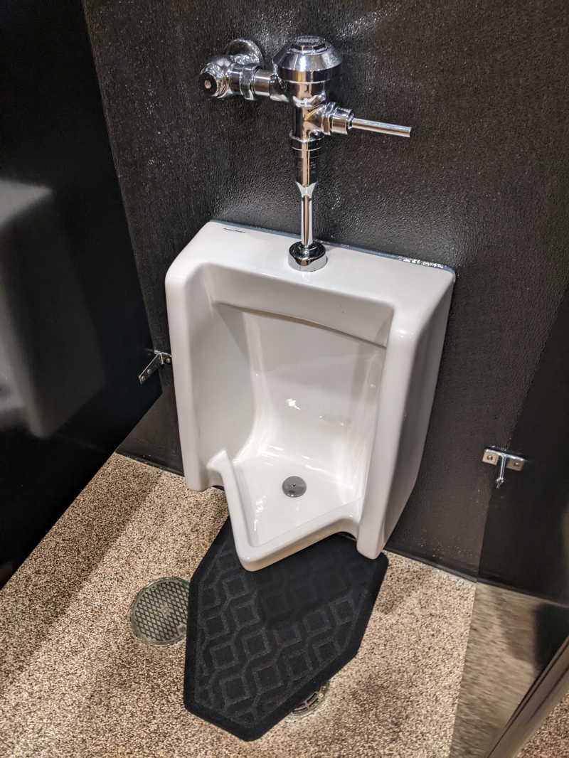 A white urinal surrounded by black walls. A black rubber mat sits underneath. Photo by Tom Ackerman.