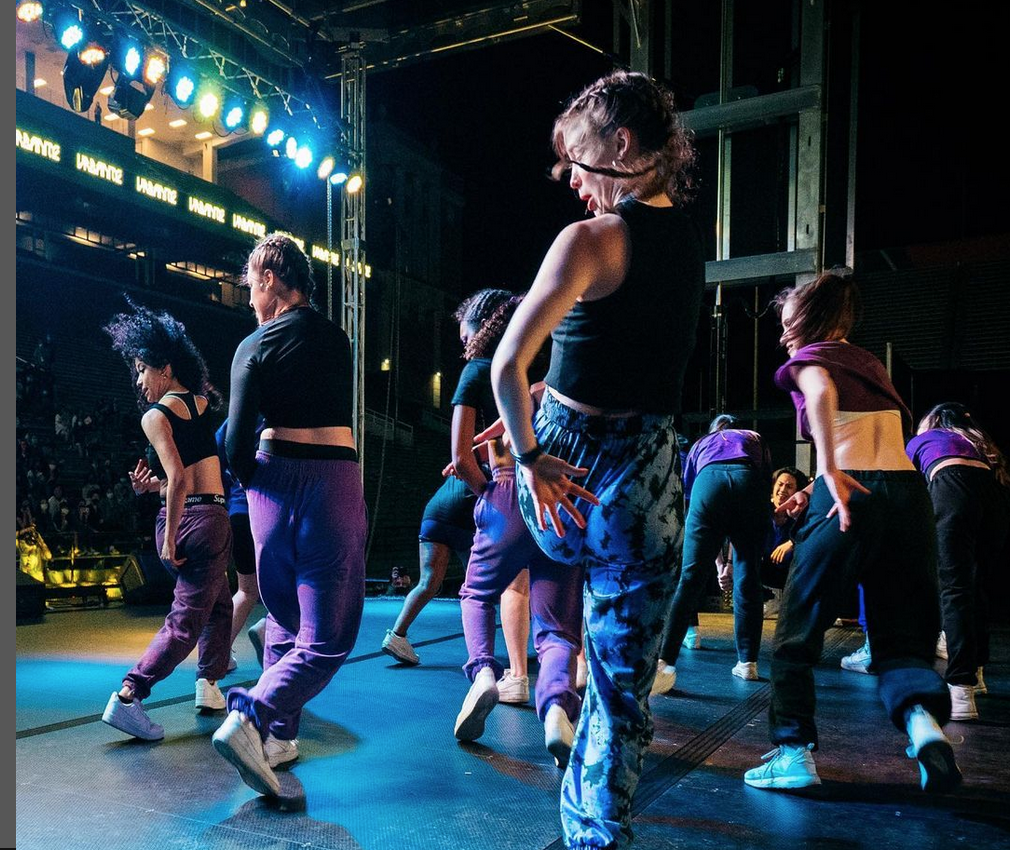 A group of dancers, mid dance, as seen from a side stage. Dancers are wearing purple and black and are mid-movement. Screen shot from the URBANITE Instagram page.   