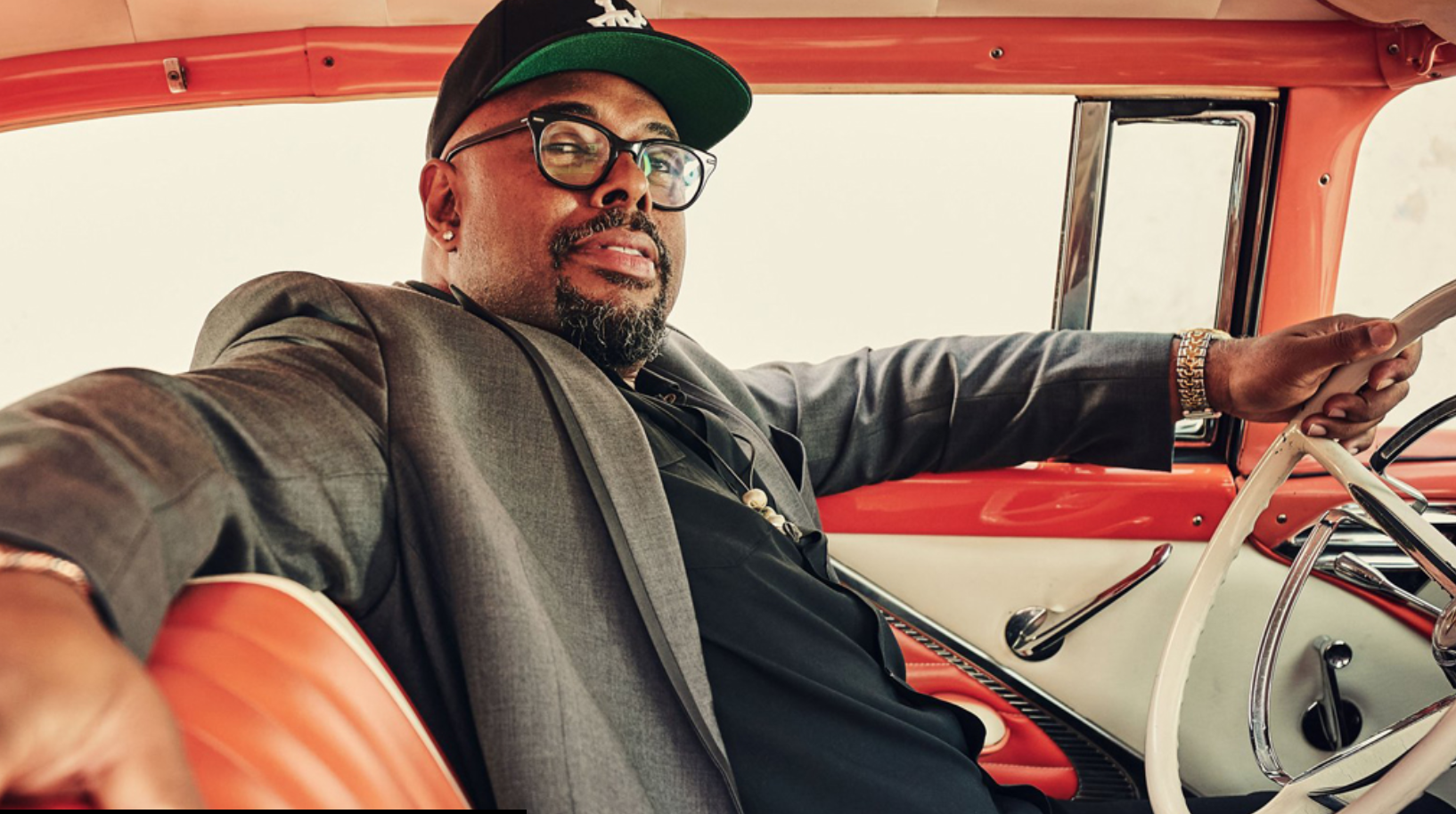 Christian McBride, a Black man in a suit jacket and a baseball cap, is seated in the driverâ€™s seat of a mid-century car. He looks to his right, toward the camera. His left hand is on the steering wheel and his right hand is slung along the back of the car seat. Screenshot from the KCPA website.