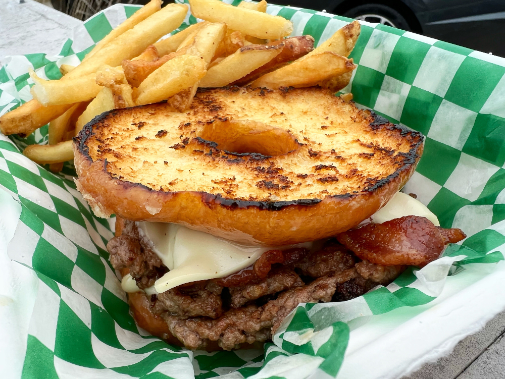 In a styrofoam container lined with green and white checkered parchment paper, there is a burger with a donut as a bun with a side of fries. Photo by Alyssa Buckley.