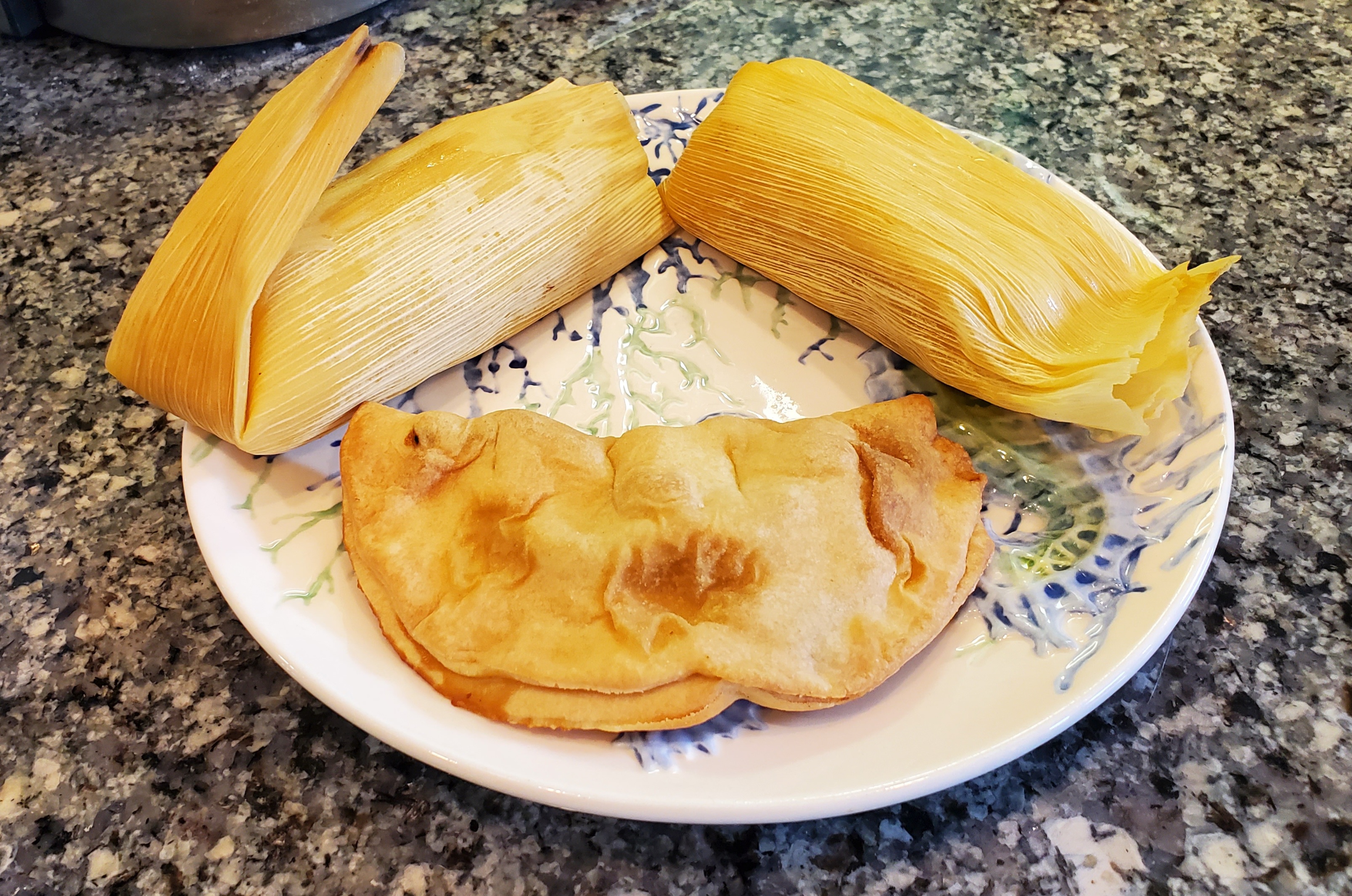 On a white plate with blue art, there are two tamales wrapped in corn husk and a soft empanada also on the table. Photo by Carl Busch.