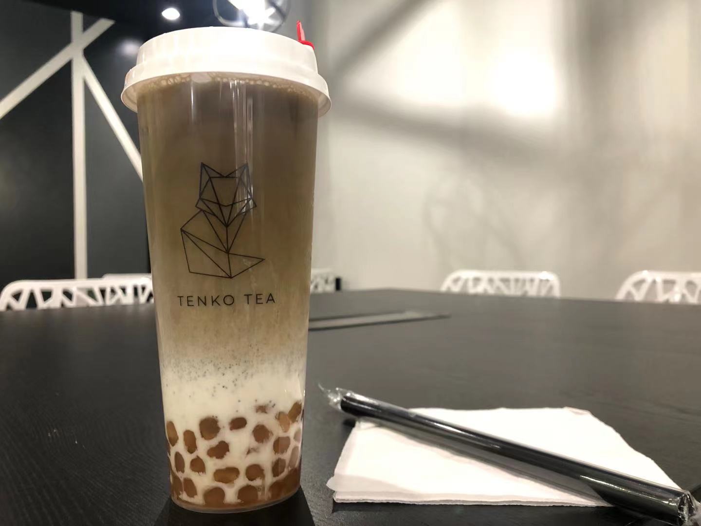 On a black table, there is a plastic cup of boba tea with Tenko Tea and the fox on the outside. Beside the cup is a white square napkin and black plastic straw wrapped in plastic. Photo by Xiaohui Zhang.
