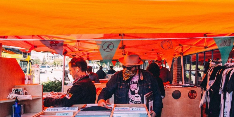 Masked outdoor shoppers flip through bins of vinyl records under an orange canopy.