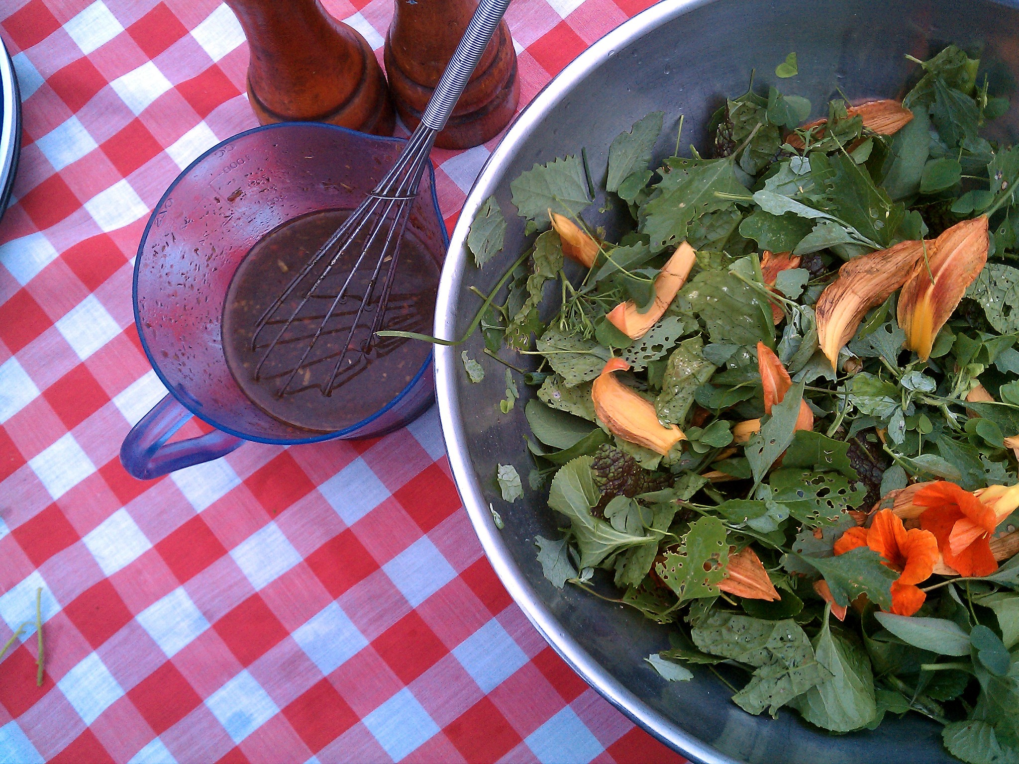 A birdâ€™s eye view of a table with a red checkered table cloth, a measuring cup with salad dressing, and a large bowl with a salad in it. The salad has edible flowers in it. Photo from the Delight Flower Farm foraging event page.
