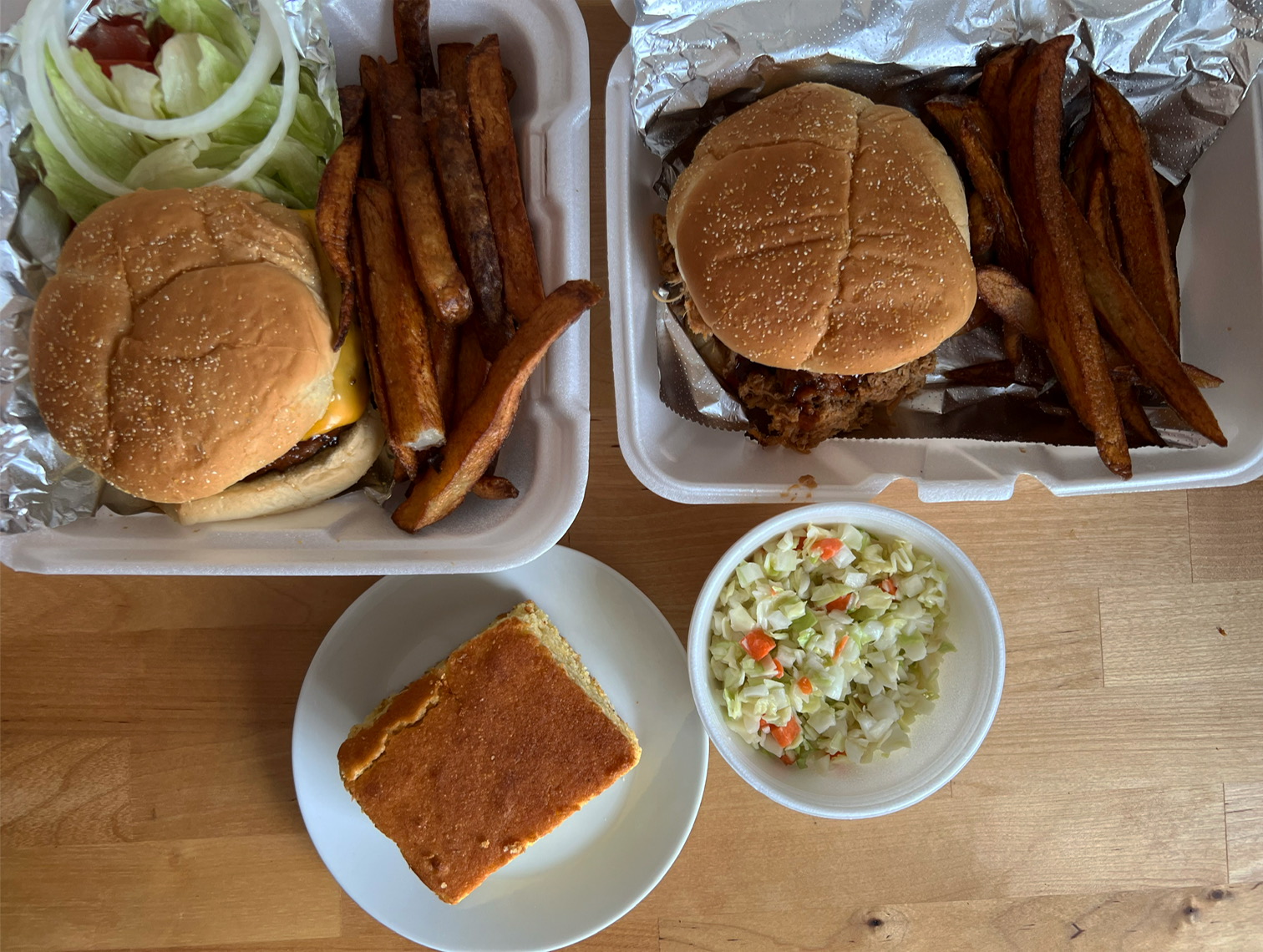 On a wooden table, there is a takeout order of burger, pulled pork, coleslaw, and cornbread. Photo by Alyssa Buckley.