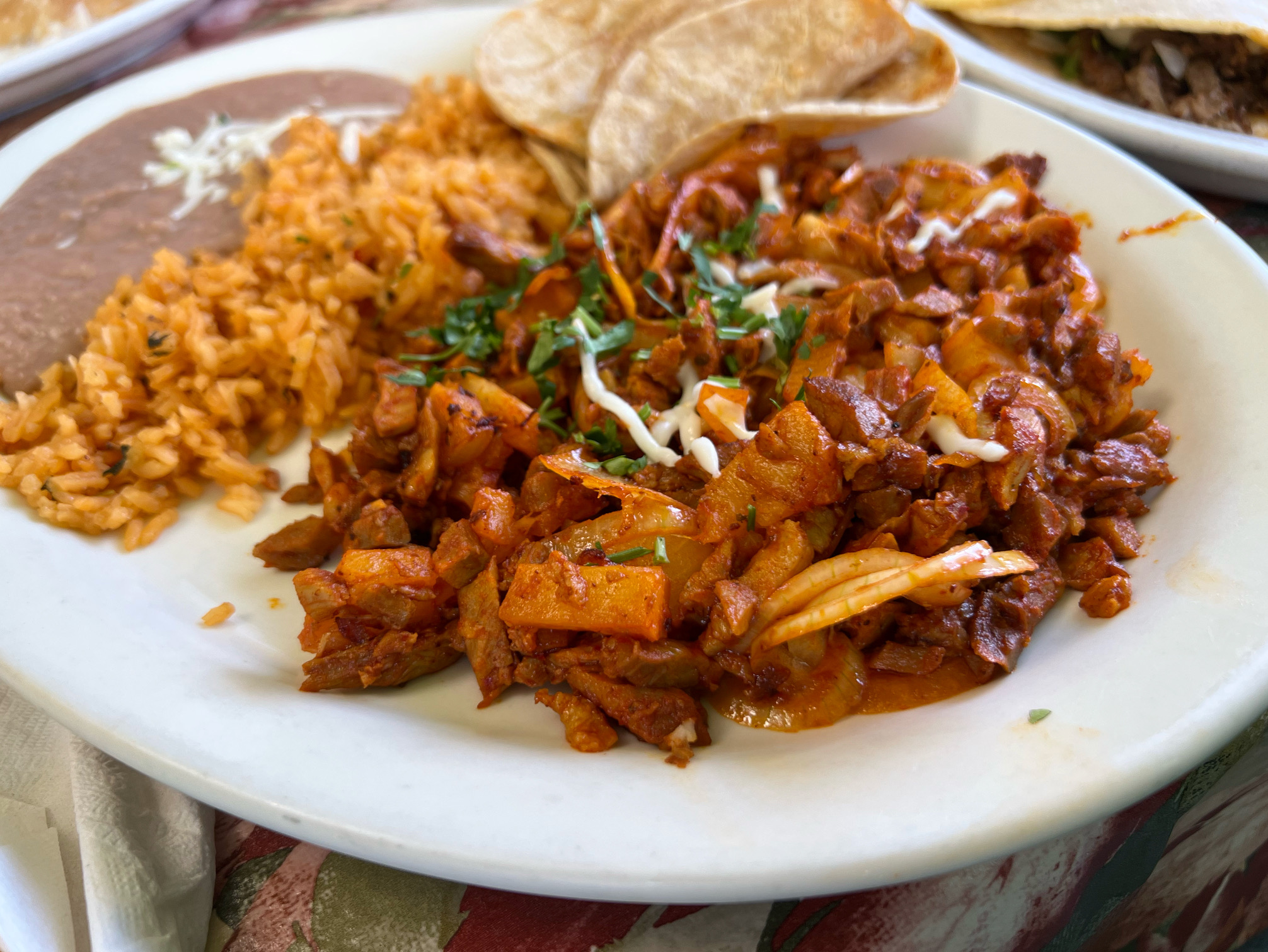 On a white plate on the iconic tablecloth of Huaraches Moroleon, there is a plate of al pastor meat with cooked onions, cilantro, and a few strings of shredded cheese. Also on the plate blurred in the background to the left is light orange rice and thin pinto beans with white shredded cheese on top. Photo by Alyssa Buckley.