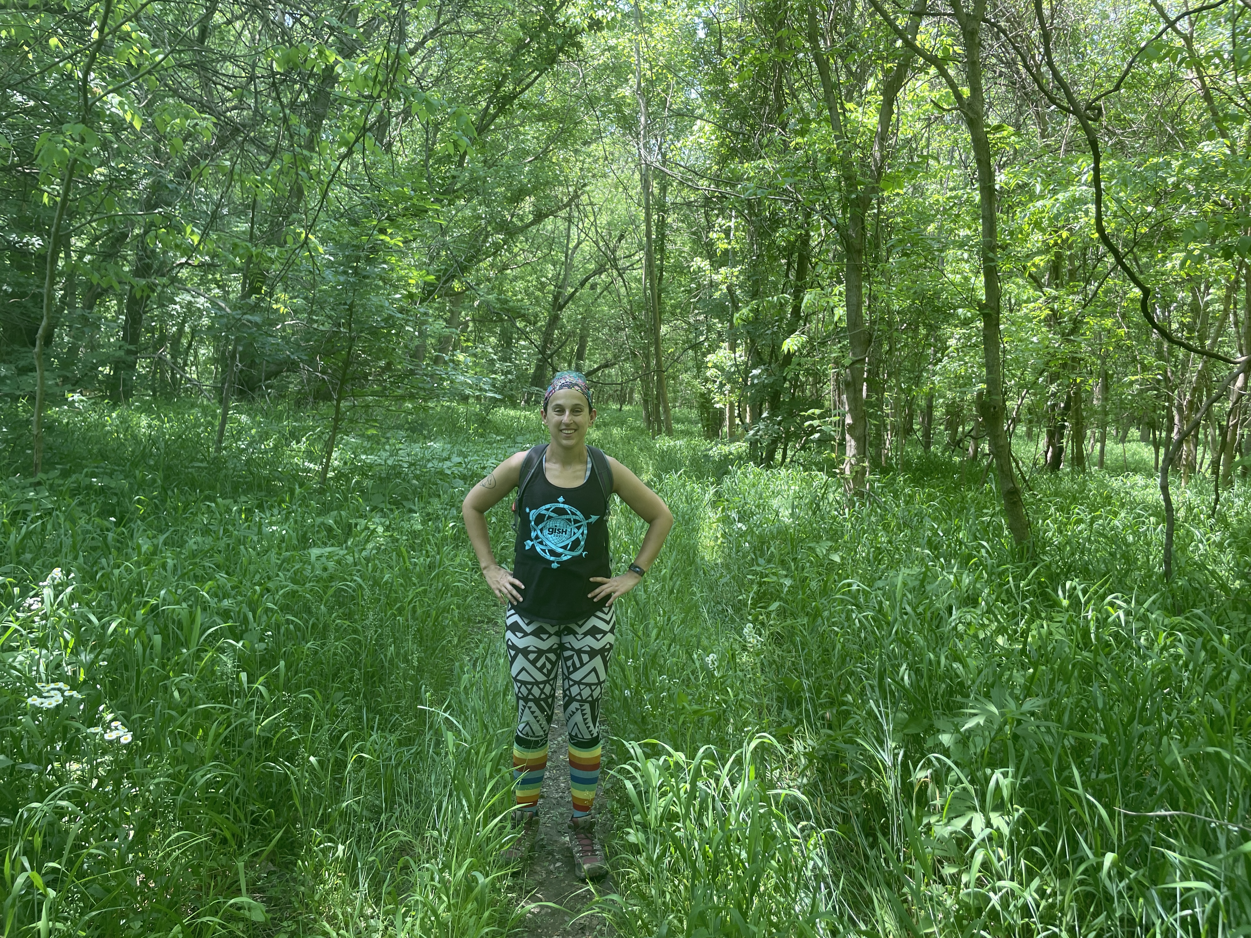 Photo of author Mara Thacker standing on a narrow trail with tall grass on either side. Mara stands with hands on hips. There are trees in the background. Main color in the photo is green.