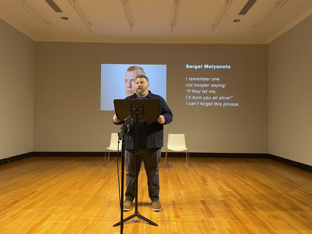 Aaron MuÃ±oz performs as part of Voices of the New Belarus at Krannert Art Musuem. He stands in front of a microphone. Behind him is a projection of the person whose words he is reading and the quote he is reading. Photo by  Landon Sinclair.