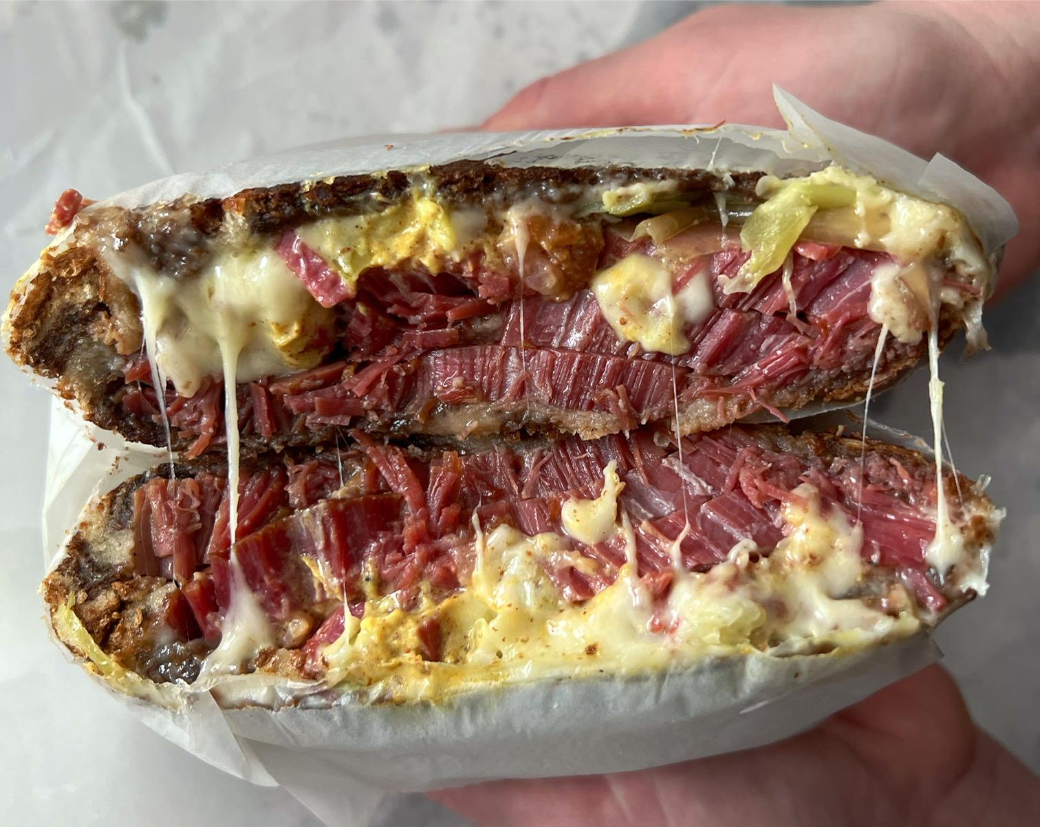 Two white hands hold open a sliced pastrami sandwich from Baldarotta's. The pastrami is red with light yellow baby Swiss cheese threads across and yellow mustard spread. Photo by Alyssa Buckley.