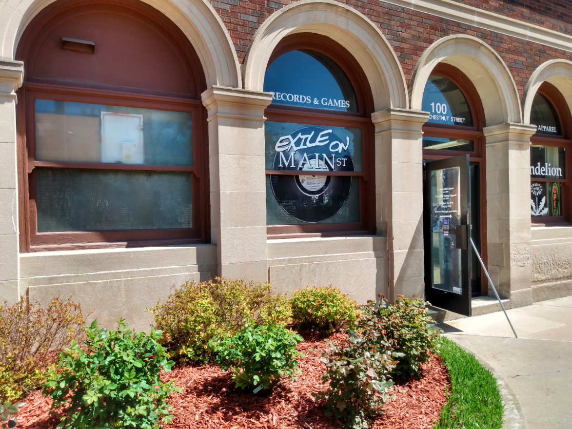 Exile on Main Street's storefront. There is green shrubbery in the foreground with a glass door propped open against a red brick and stone facade. The leftmost window display's the store name with an image of a record, and the rightmost window shows the logo to Dandelion (the store they share a space with).