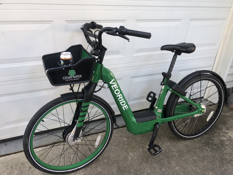 A green electric bike with a black basket hanging on the front sits in front of a white garage door. It has the word Veoride in white lettering on the body. Photo by Andrea Black.