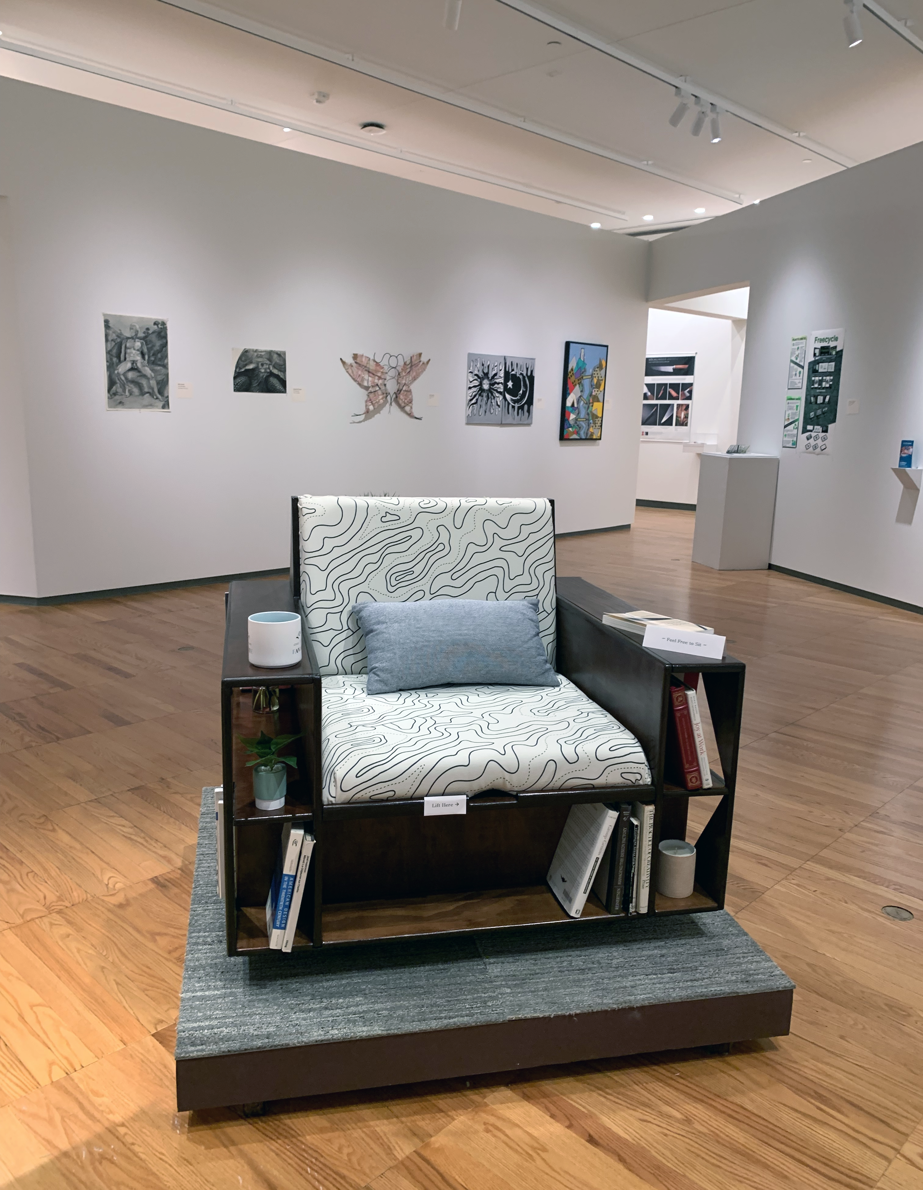 Chair built with arms that hold shelves in the foreground with other paintings, sculptures, and drawings on the back wall.