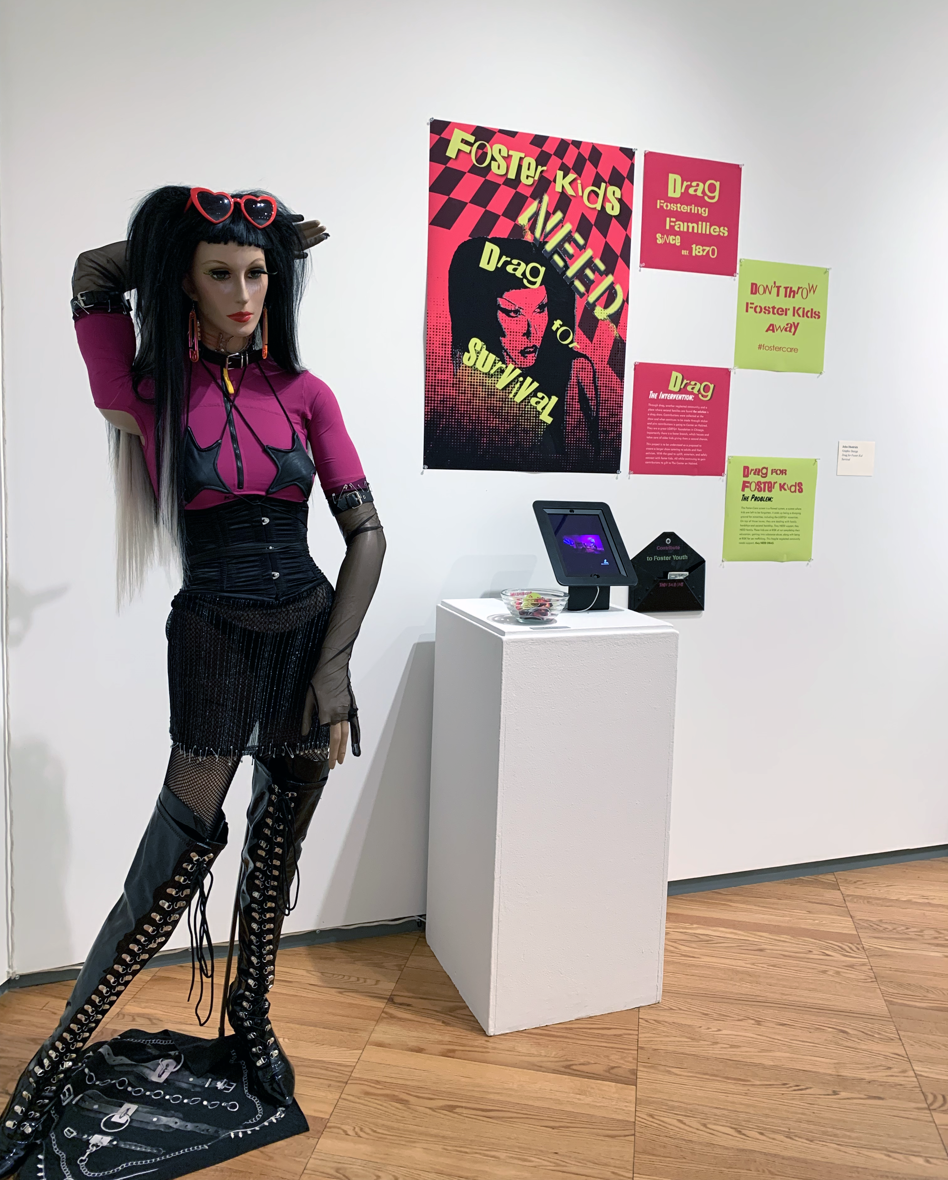 Maniken in drag in front of posters and digital displays for Drag for Foster Kids project.