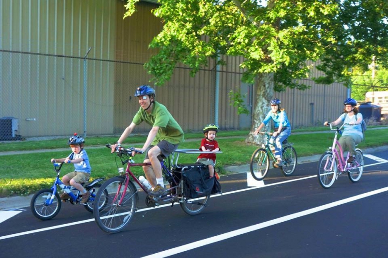 A group bicycling along the side of the road. They are led by a young boy on small blue bike, followed by a dad on a maroon bike with a small child on a seat behind him. Then there are two women behind them. Photo from Facebook event page.