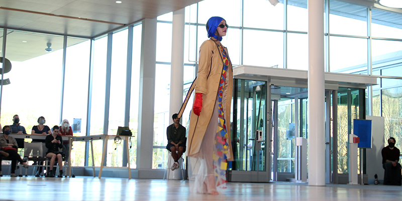 Female model wearing long tan trench over multicolored dress and purple headscarf.