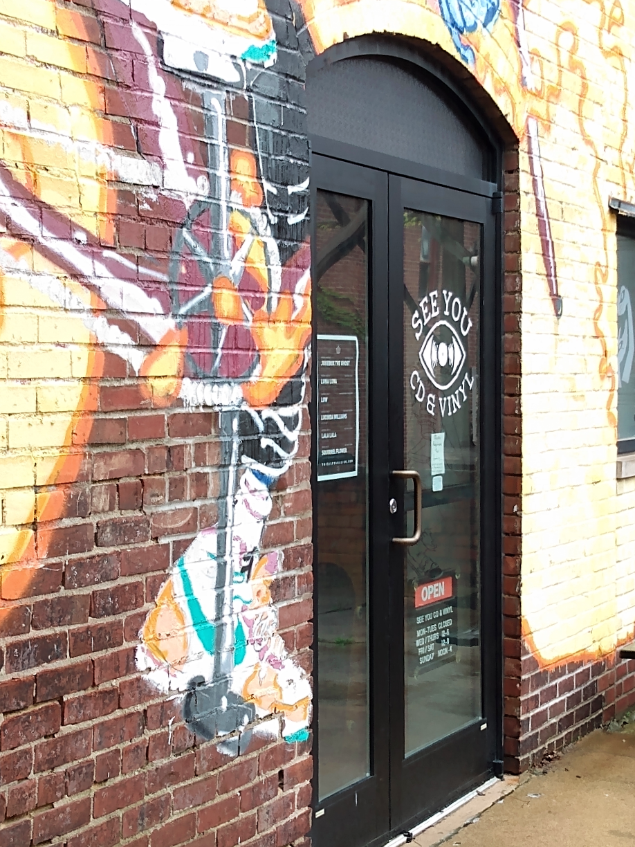 The entrance to See You CD & Vinyl from the back alley. The brick wall has been painted with graffiti-style art, and a glass door displays the store logo: a stylized eyeball with a record for the pupil. A poster for upcoming shows from Pygmalion the store's hours are on display.