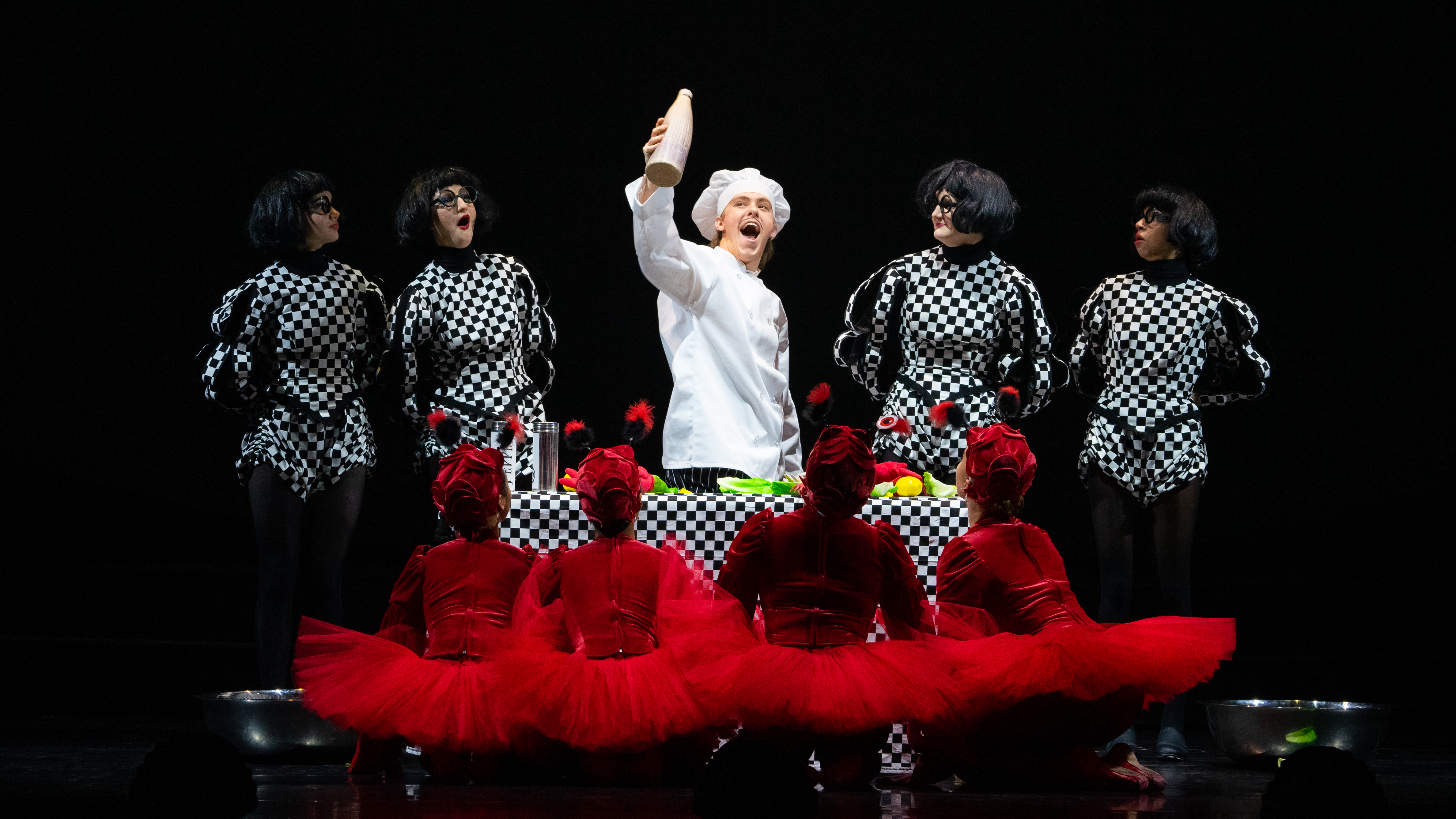 Onstage at the Virginia Theatre, a chef holds a bottle high in the air. Beside the chef, four minions in black and white checkered shirts stand beside him, two on each side. In front, facing away from the camera, four crabs in red tutus face the chef in CU Ballet's The Little Mermaid. Photo by Claire Daly.