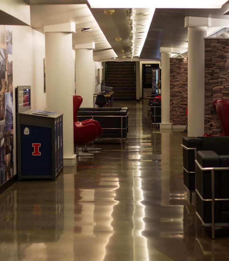 A hallway with cylindrical columns along either side. In the foreground is a blue recycling bin with an orange block I. There are black leather chairs with metal legs along the sides. Photo by Jorge Murga.